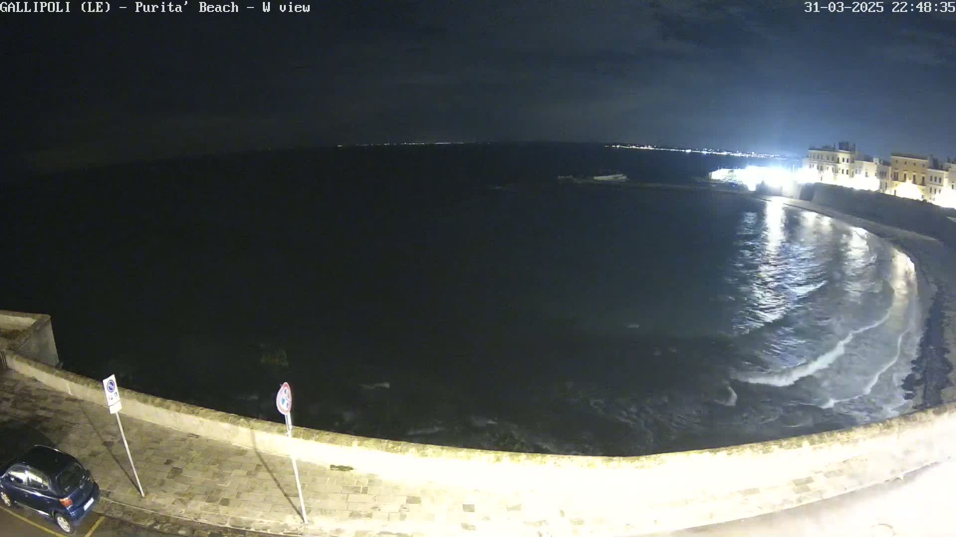 A nighttime view of a calm, dark sea gently lapping against a stone wall, with a few lights visible in the distance.