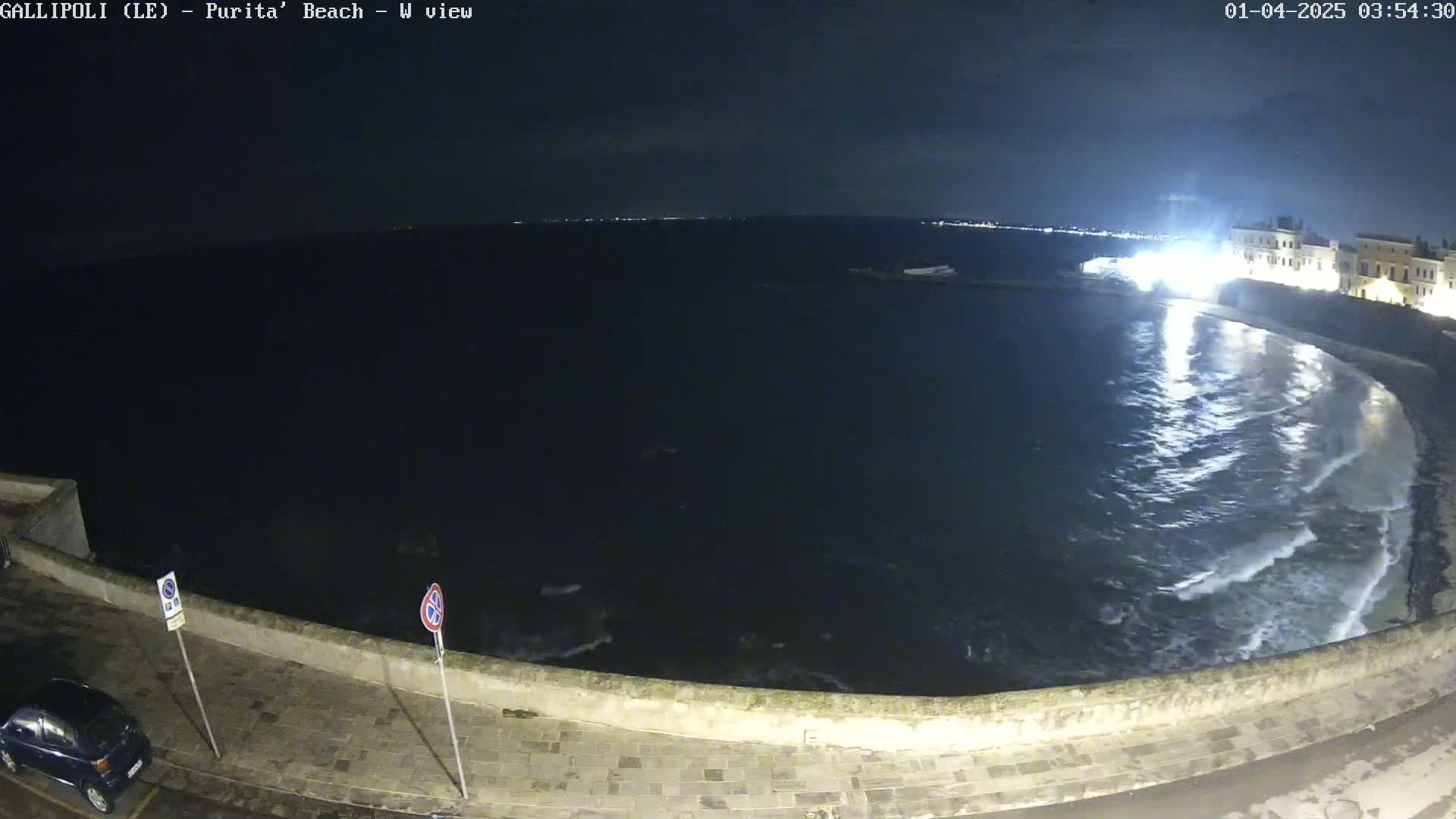 A nighttime view of a calm, dark sea lapping gently at a stone wall, with a few lights visible in the distance.