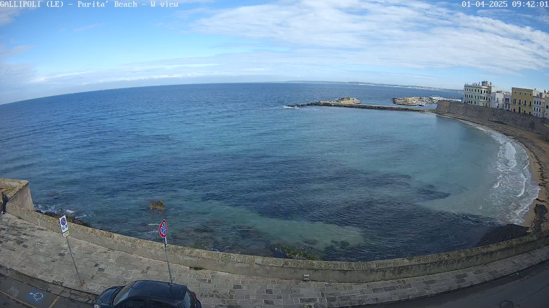 A coastal scene on a partly sunny day shows a calm sea curving around a stone wall, next to which a paved road leads to a small sandy beach with gentle waves.