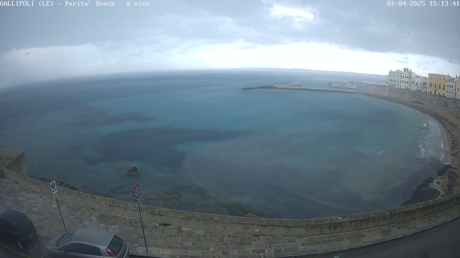 A partly cloudy day overlooks a calm bay with a stone wall and buildings along its edge, and parked cars visible in the foreground.