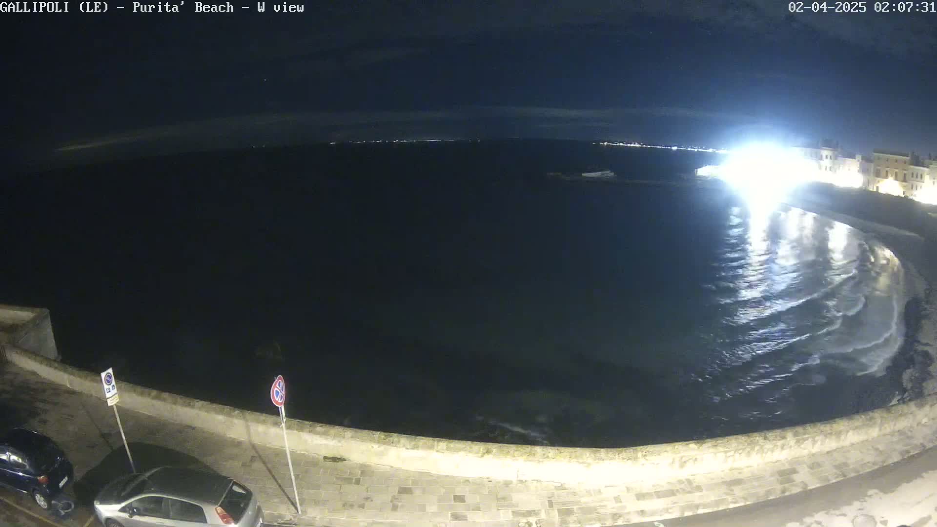 A nighttime view of a dark, wavy ocean meeting a stone wall along a shore, with a few parked cars visible on the land.
