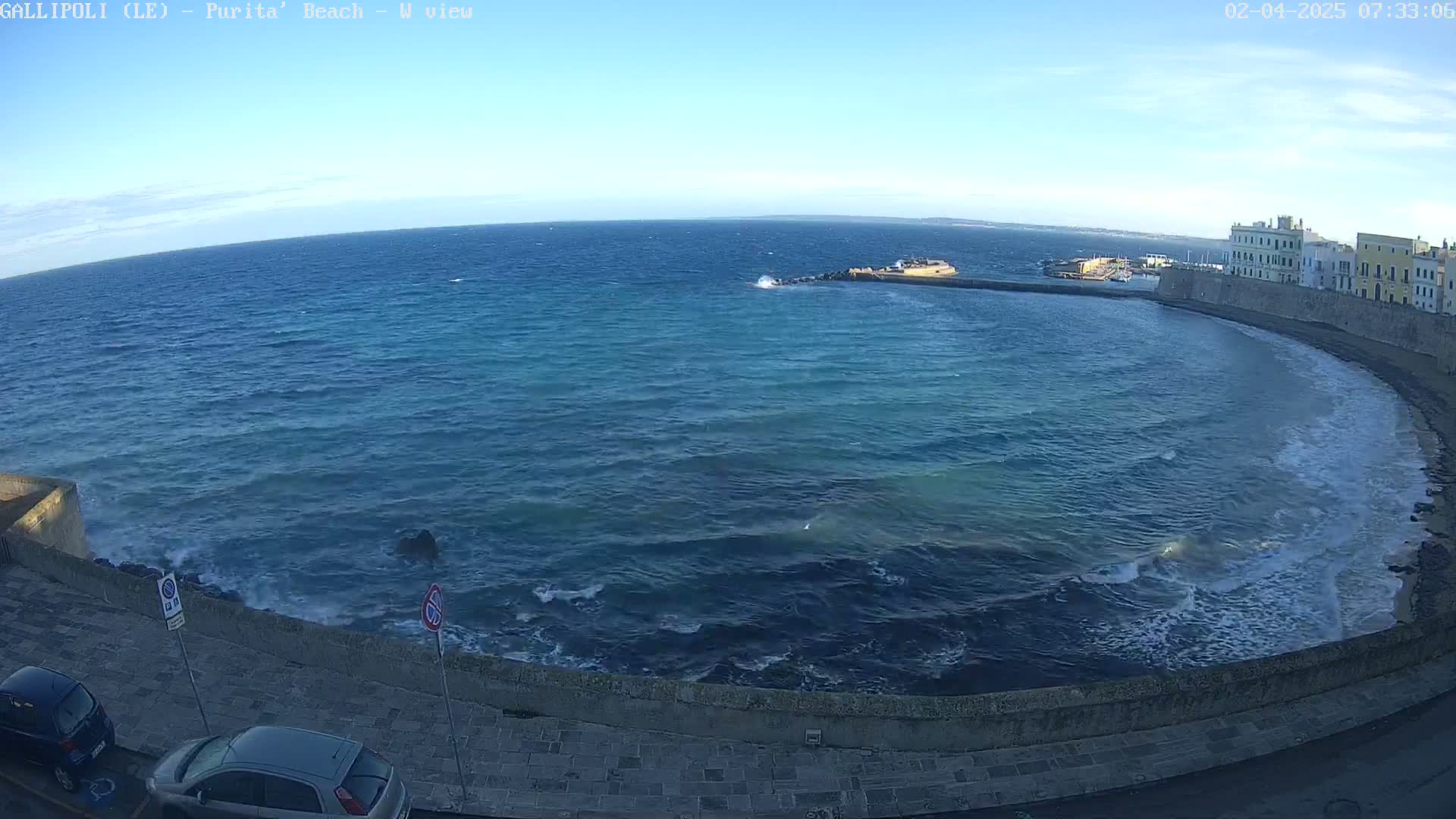 A clear, sunny day reveals a coastal scene with a calm sea lapping gently at a stone wall, and a row of buildings visible in the distance.