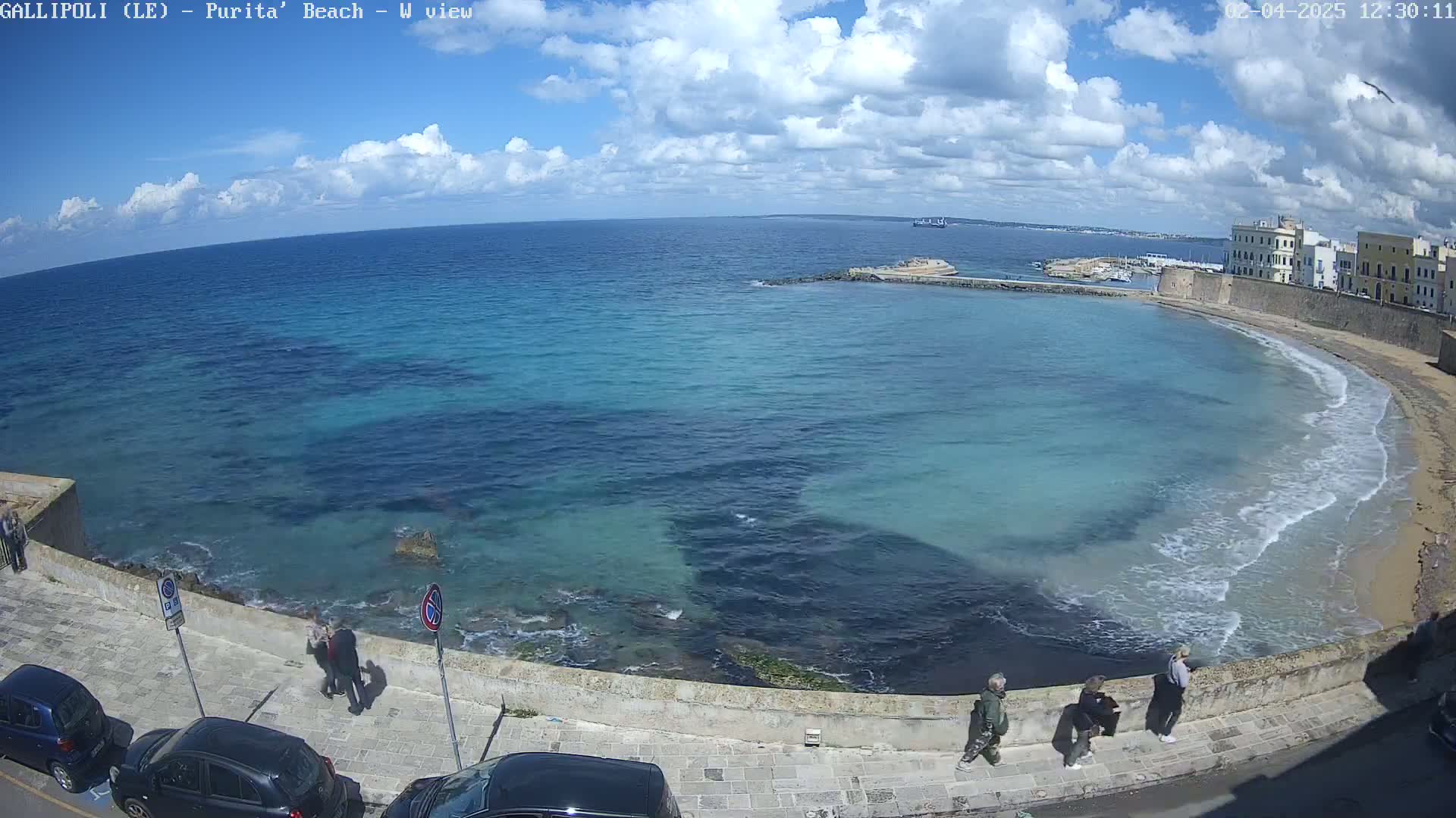 A partly sunny day shows a partially enclosed bay with turquoise water lapping a sandy beach next to a stone wall and buildings; several people are standing on a paved walkway overlooking the water.