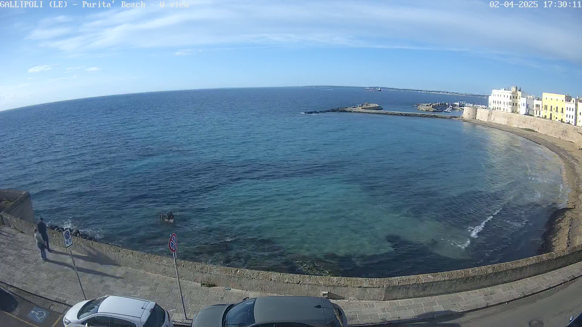A clear, sunny day overlooks a calm, teal-colored bay with a stone wall and buildings along the shore, and two cars parked nearby.