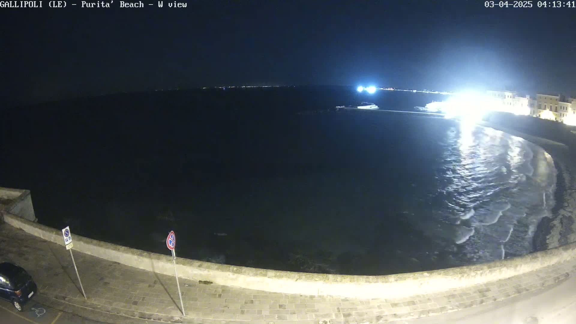 A nighttime view of a dark, wavy ocean lapping a shoreline with a low stone wall and a car parked nearby.