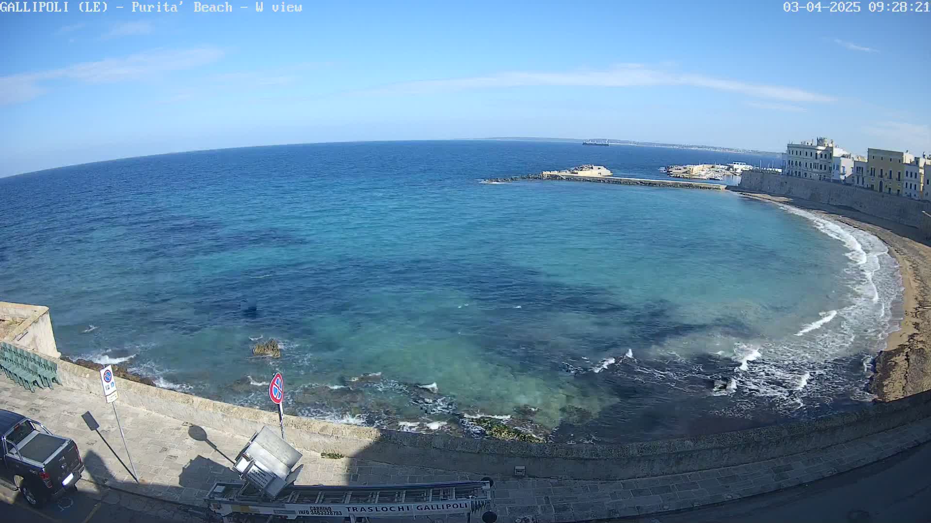 A clear, sunny day shows a curving beach with turquoise water lapping against a rocky shore and a town in the distance.