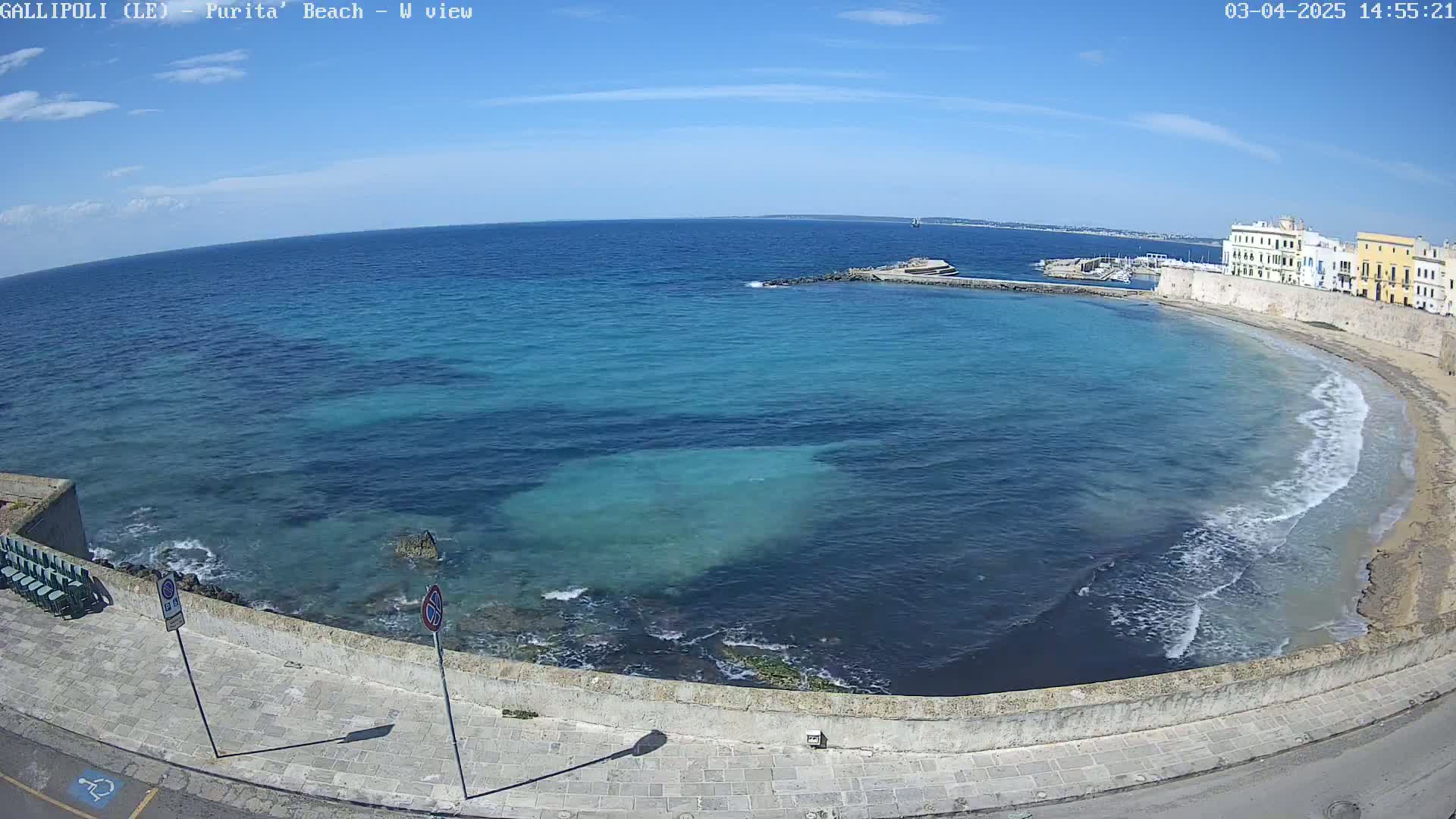 A clear, sunny day shows a curving shoreline with a light-colored sandy beach, turquoise water lapping gently at the shore, and buildings behind a low wall.