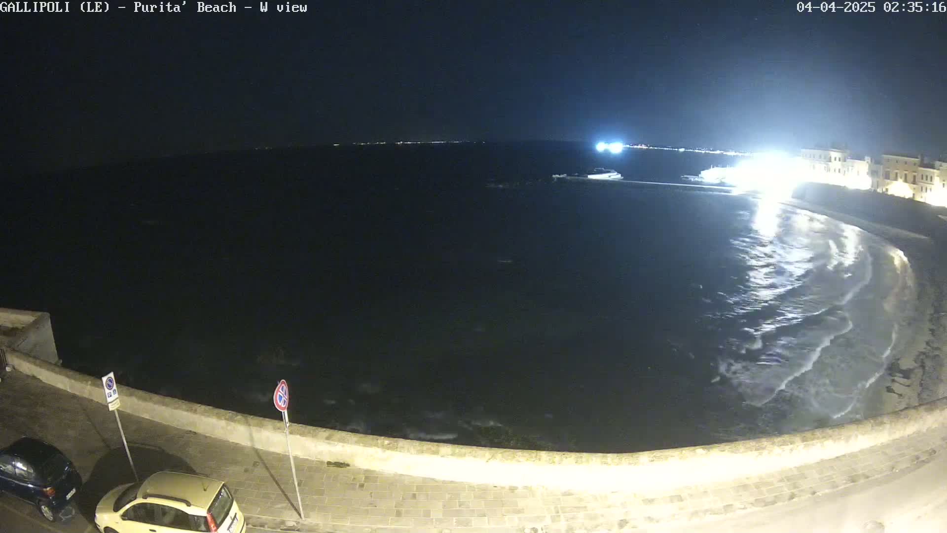 A nighttime view of a dark, wavy sea lapping a crescent-shaped beach next to a stone wall and street with parked cars.