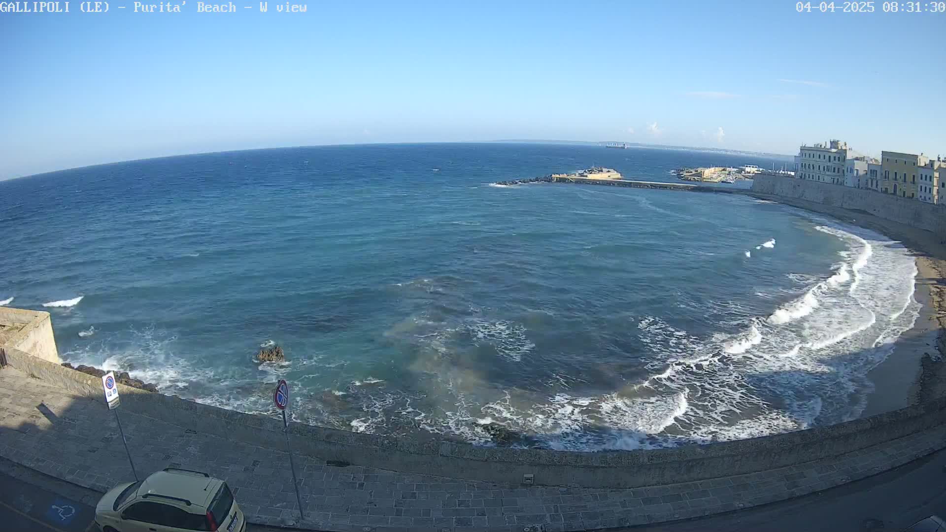 A clear, sunny day shows a coastal view of a city wall and beach with gentle waves lapping the shore.