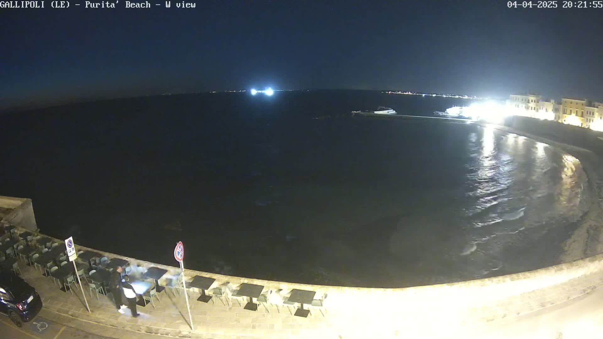 A nighttime, seaside view shows a calm, dark body of water lapping gently against a stone wall lined with outdoor seating, with two people walking along the wall.