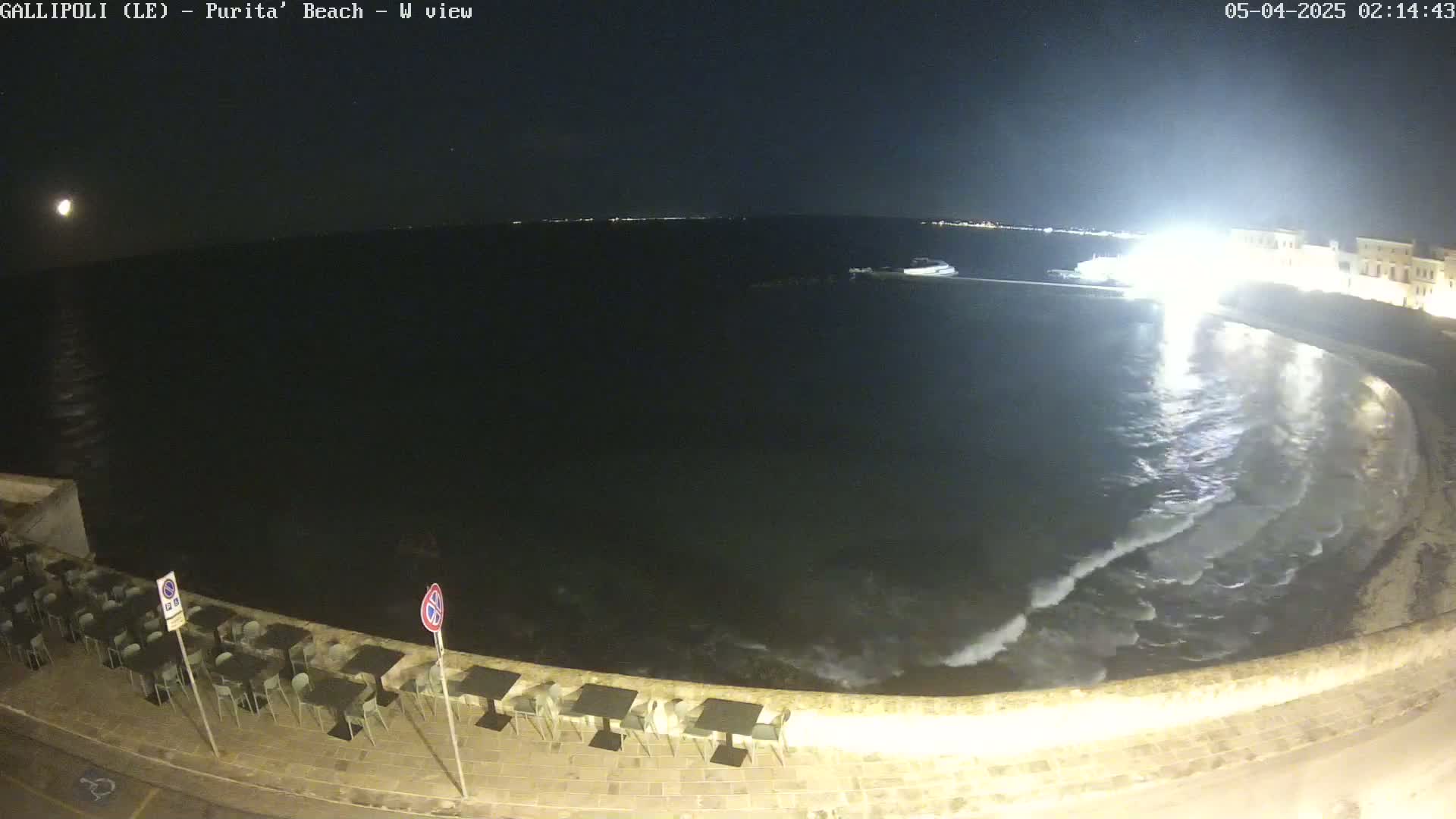 A nighttime view of a dark, wavy ocean lapping against a stone shoreline with empty tables and chairs at an outdoor restaurant under a clear sky.