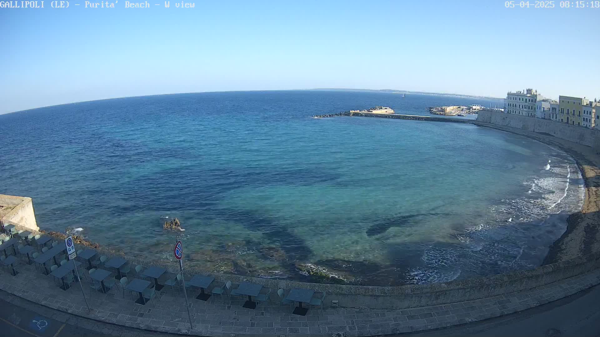 A clear, sunny day reveals a calm bay with clear, turquoise water lapping a stone shoreline lined with outdoor seating.