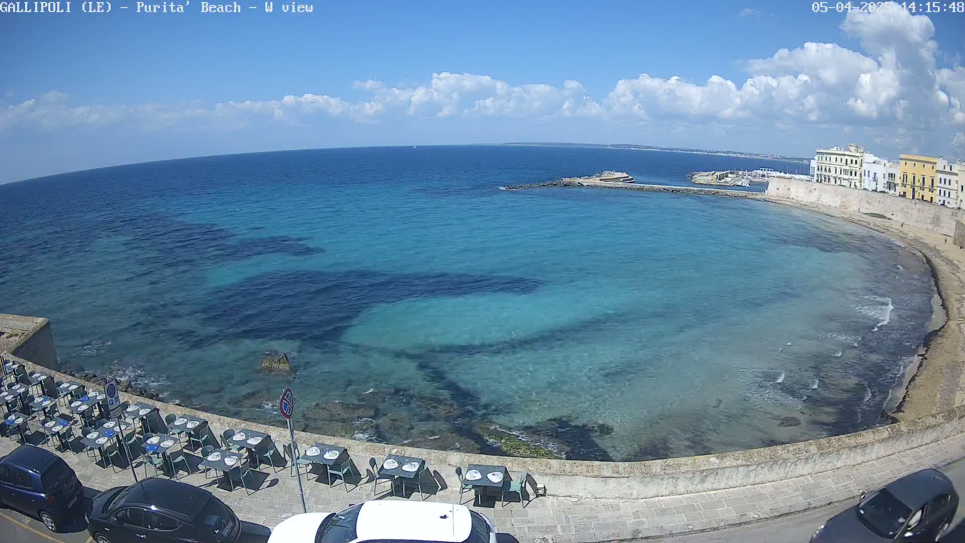 A partly cloudy day shows a clear, turquoise bay next to a stone wall lined with outdoor restaurant seating and parked cars, with buildings visible in the background.
