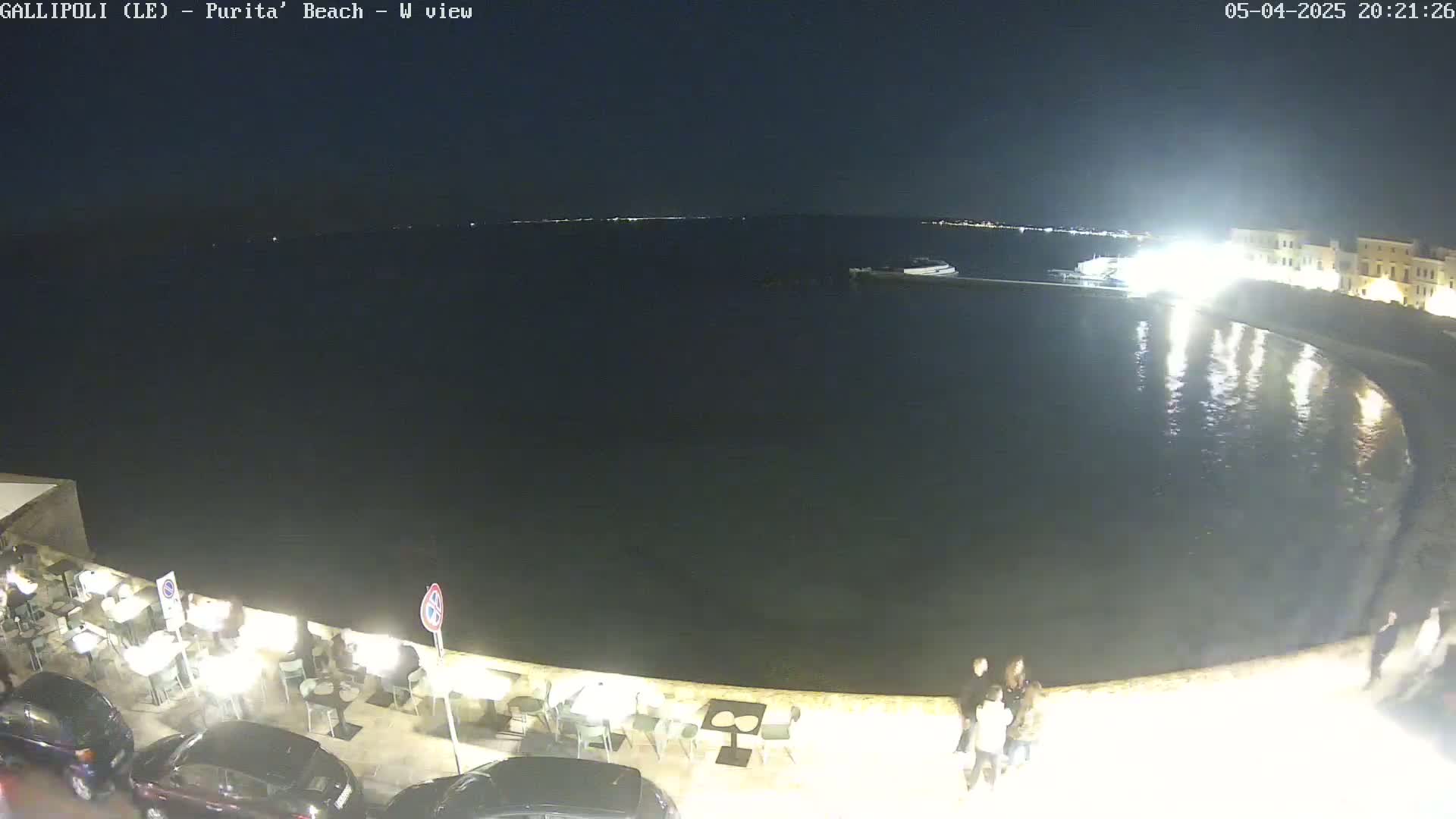 A nighttime view of a calm, dark body of water next to a waterfront restaurant with patrons seated at outdoor tables, and cars parked nearby.