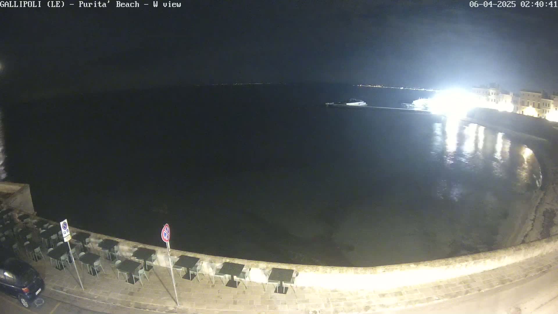 A nighttime view of a calm, dark body of water next to a stone wall lined with empty outdoor tables and chairs, under a clear sky.