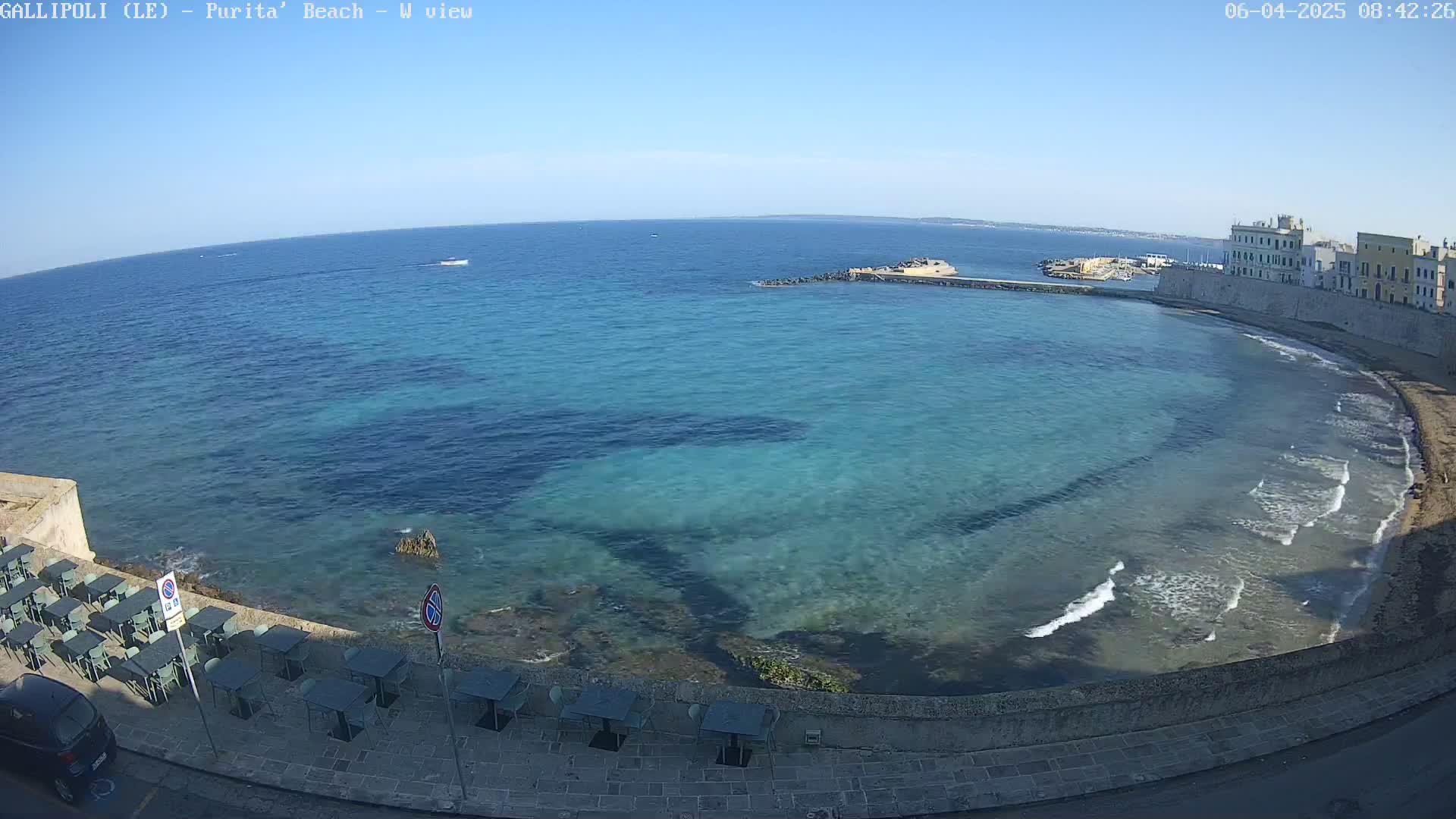 A clear, sunny day shows a turquoise-blue bay with small waves lapping a stone shore lined with outdoor cafe seating, and buildings in the distance.