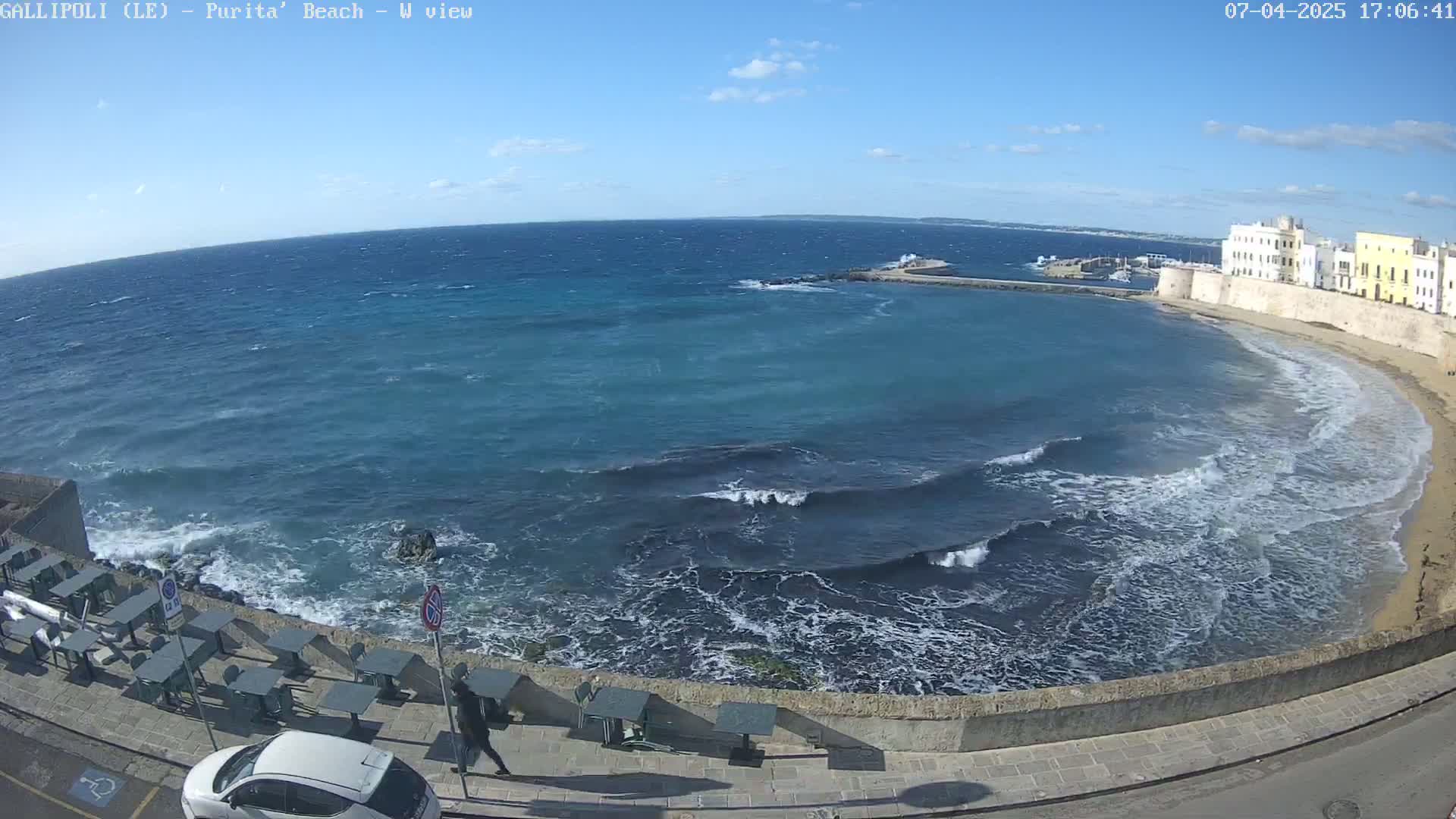 A wavy ocean meets a sandy beach along a stone wall lined with outdoor seating under a mostly clear, sunny sky.