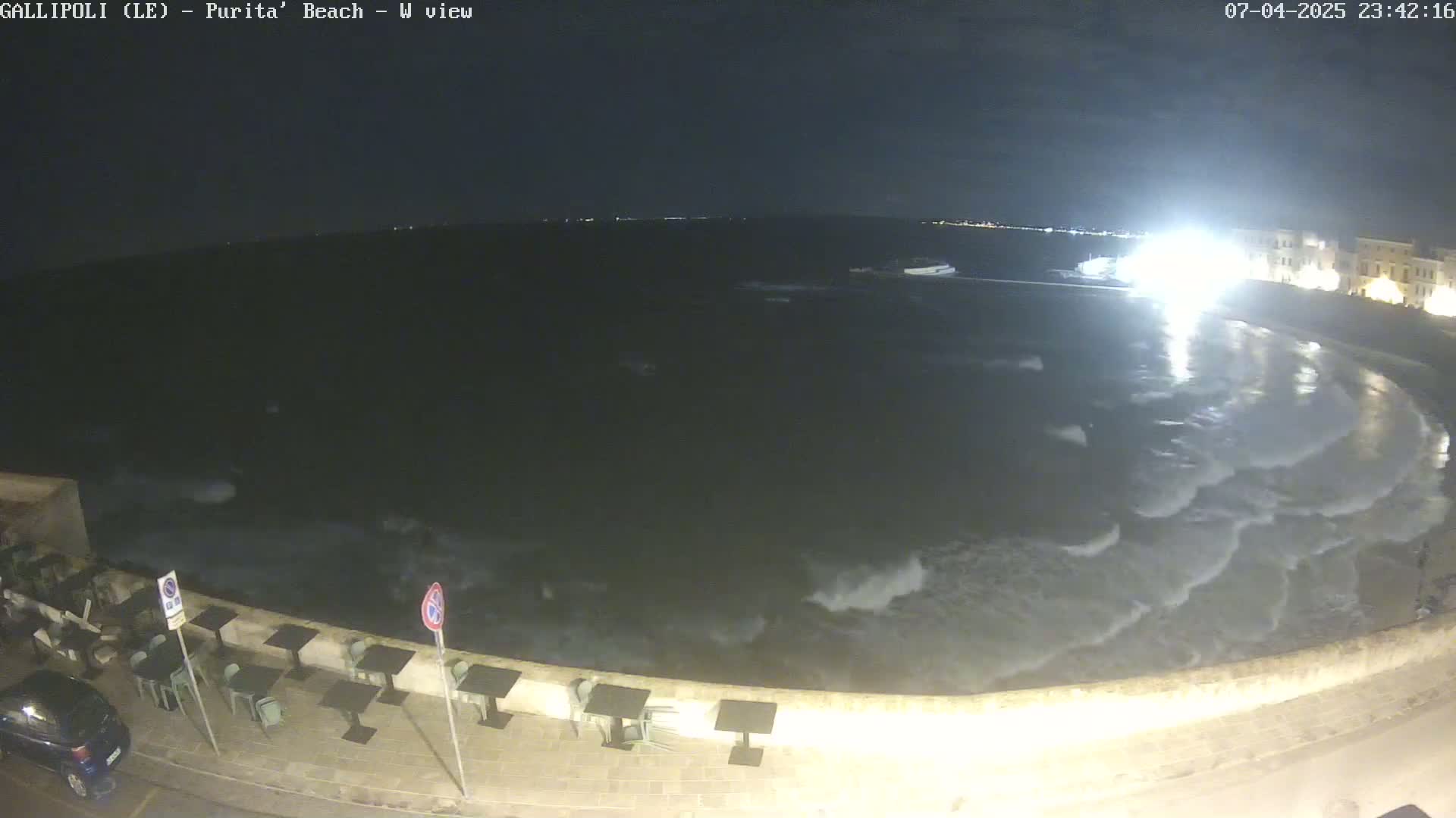 A nighttime view of a beach with gentle waves lapping the shore, some outdoor seating visible along the waterfront.
