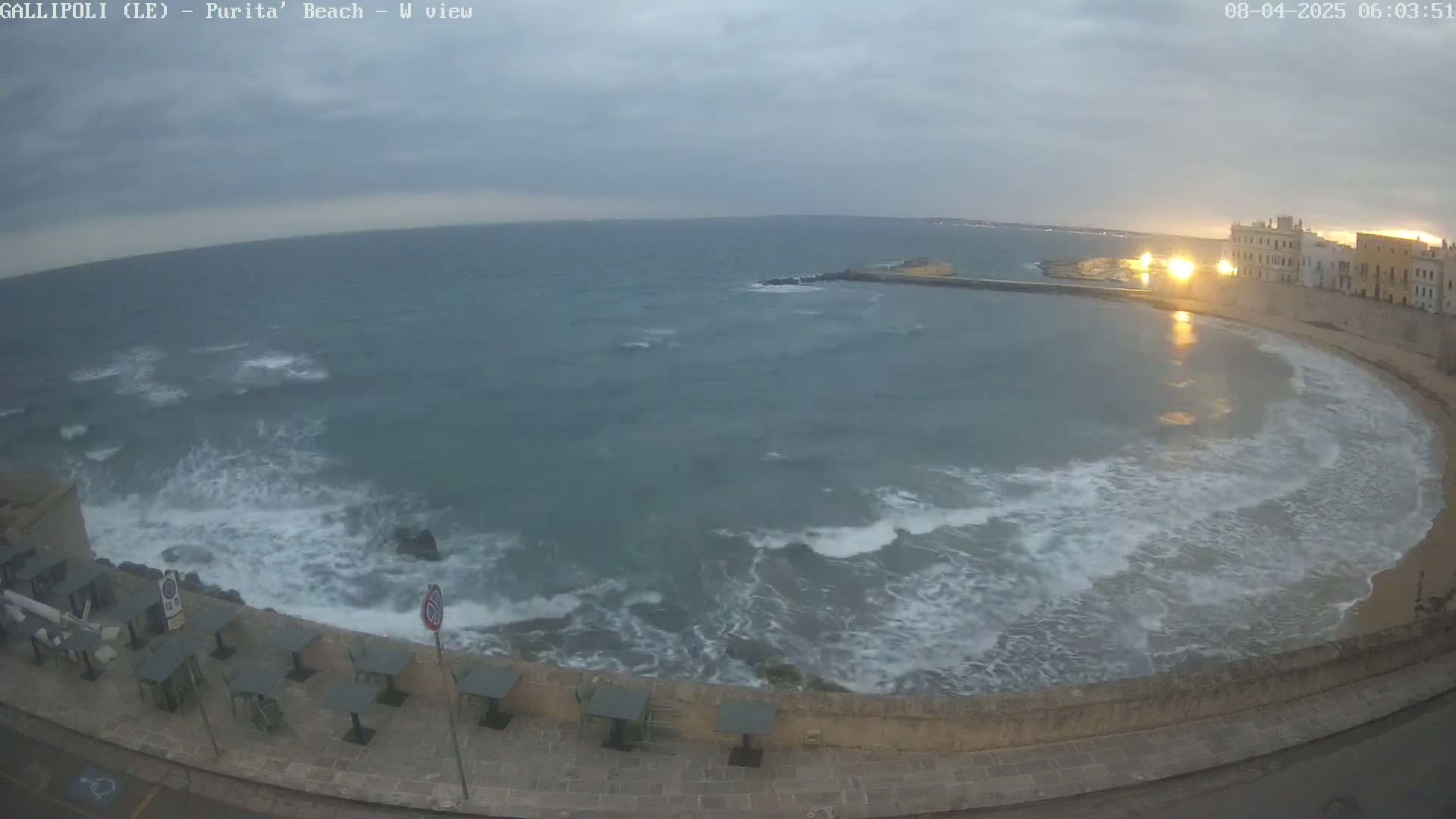 A coastal town's curved waterfront, with a calm, partially-sunlit bay and moderate waves crashing against a stone wall bordering outdoor seating under an overcast sky.