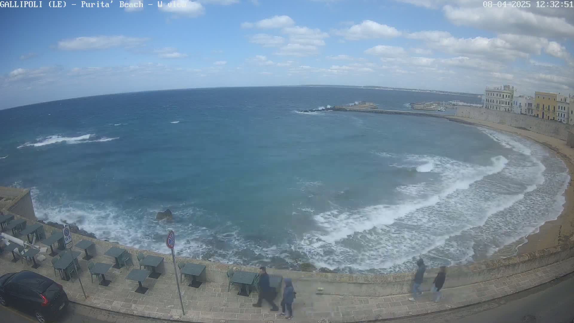 A partly cloudy day shows a wavy ocean lapping a sandy beach next to a stone wall and a row of outdoor cafe tables.