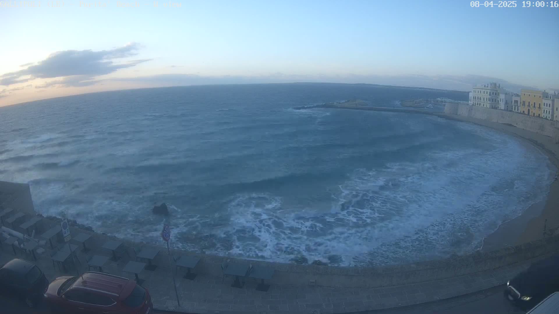 A windy, twilight view shows a choppy sea lapping against a shoreline with buildings and parked cars.