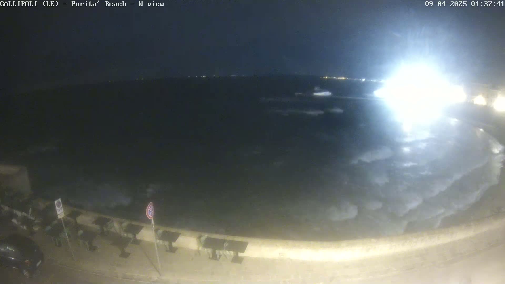 A nighttime view of a dark, somewhat choppy sea with a bright light source in the distance, and a paved area with outdoor seating in the foreground.