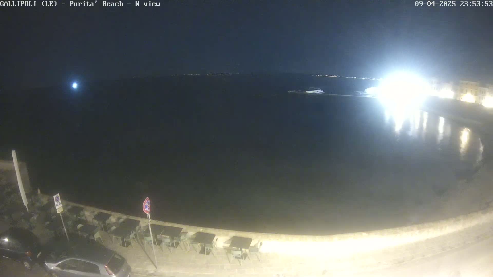 A nighttime view of a dark body of water next to a shoreline with empty outdoor seating and parked cars under a clear sky.