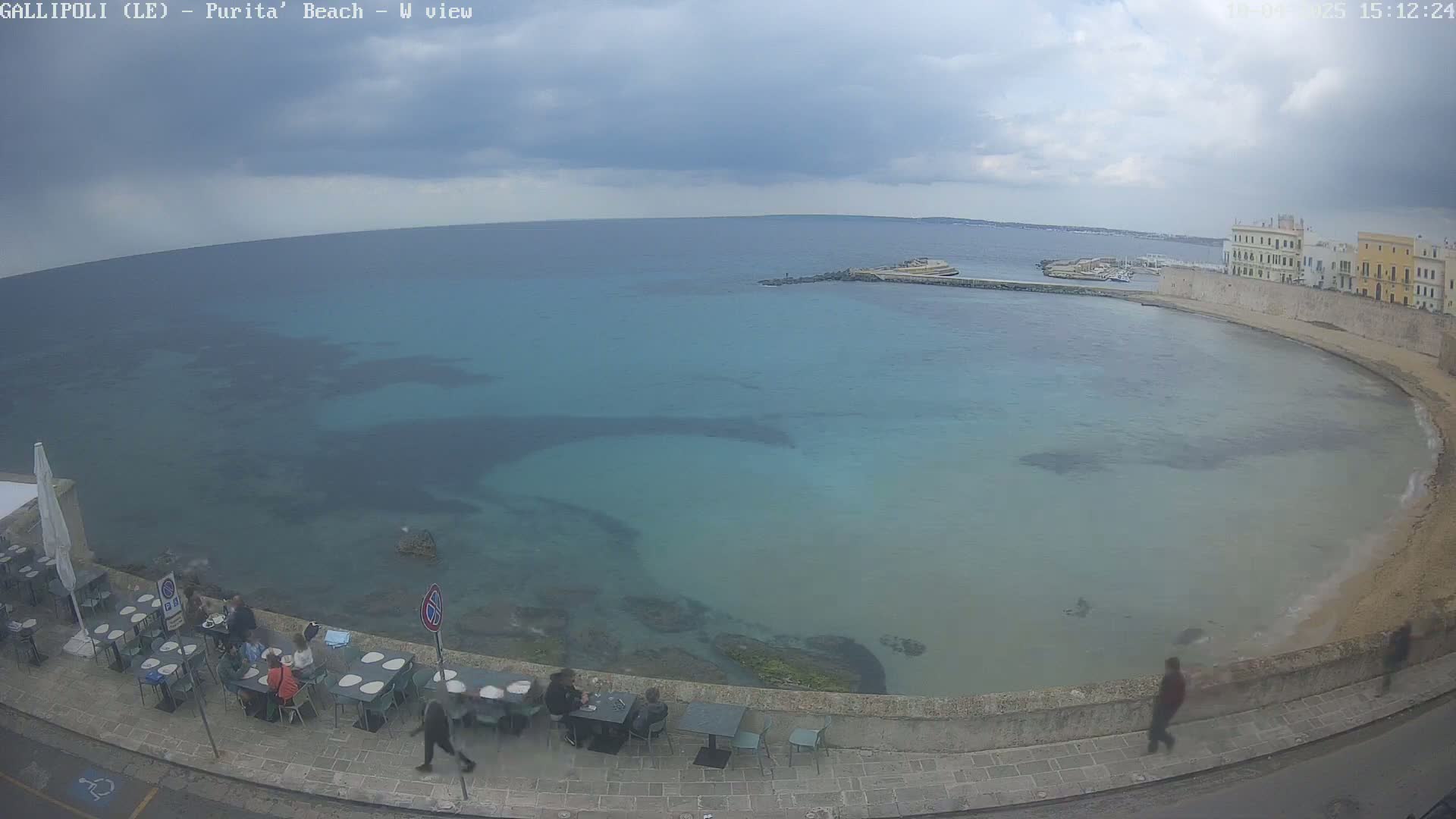A mostly cloudy day overlooks a calm, teal-colored bay with a stone wall and buildings, where several people are sitting at outdoor tables.