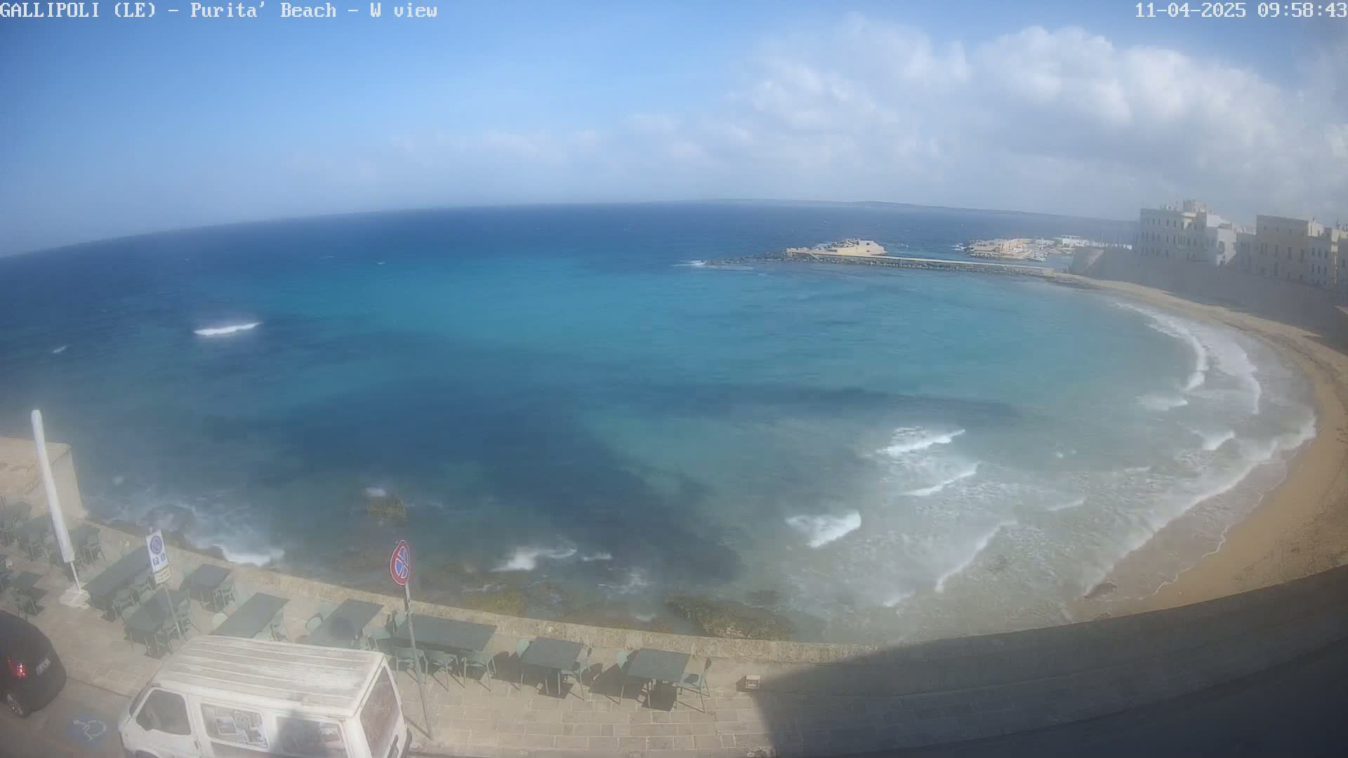 A partly cloudy day shows a view of a sandy beach curving around a calm, turquoise bay next to buildings, with small waves lapping the shore and outdoor seating visible along a paved walkway.