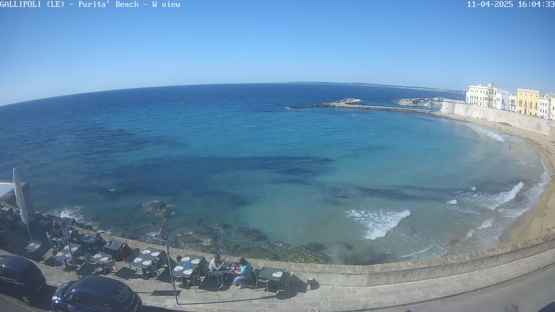 A clear, sunny day shows a picturesque beach with turquoise water lapping a sandy shore, a stone wall with buildings behind it, and a few people sitting at outdoor tables near the water's edge.