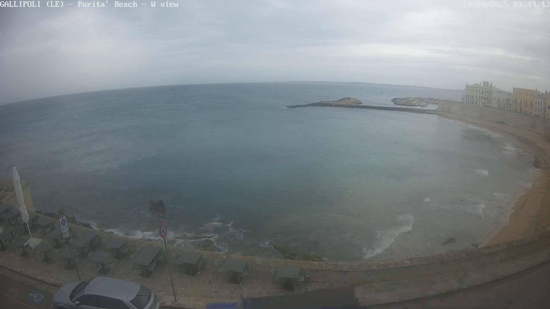 A partly cloudy day shows a calm sea curving into a sandy beach next to stone buildings and outdoor seating.