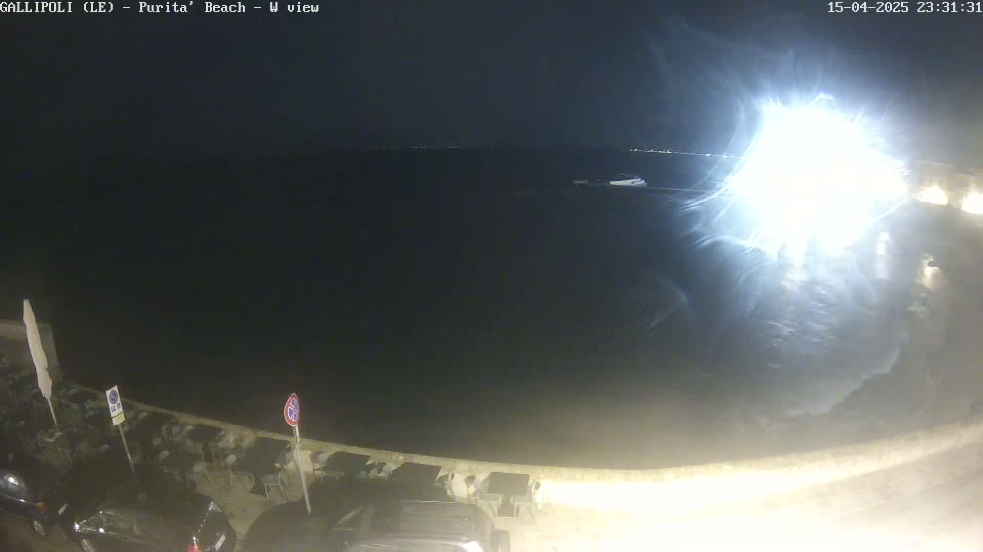 A nighttime, seaside view shows a dark body of water reflecting a bright light source, with parked cars and outdoor furniture visible along a shoreline.