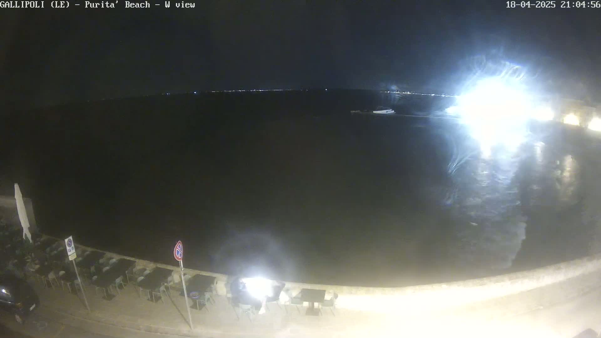 A nighttime view of a dark body of water, a shoreline with empty outdoor seating, and a bright light source in the distance.