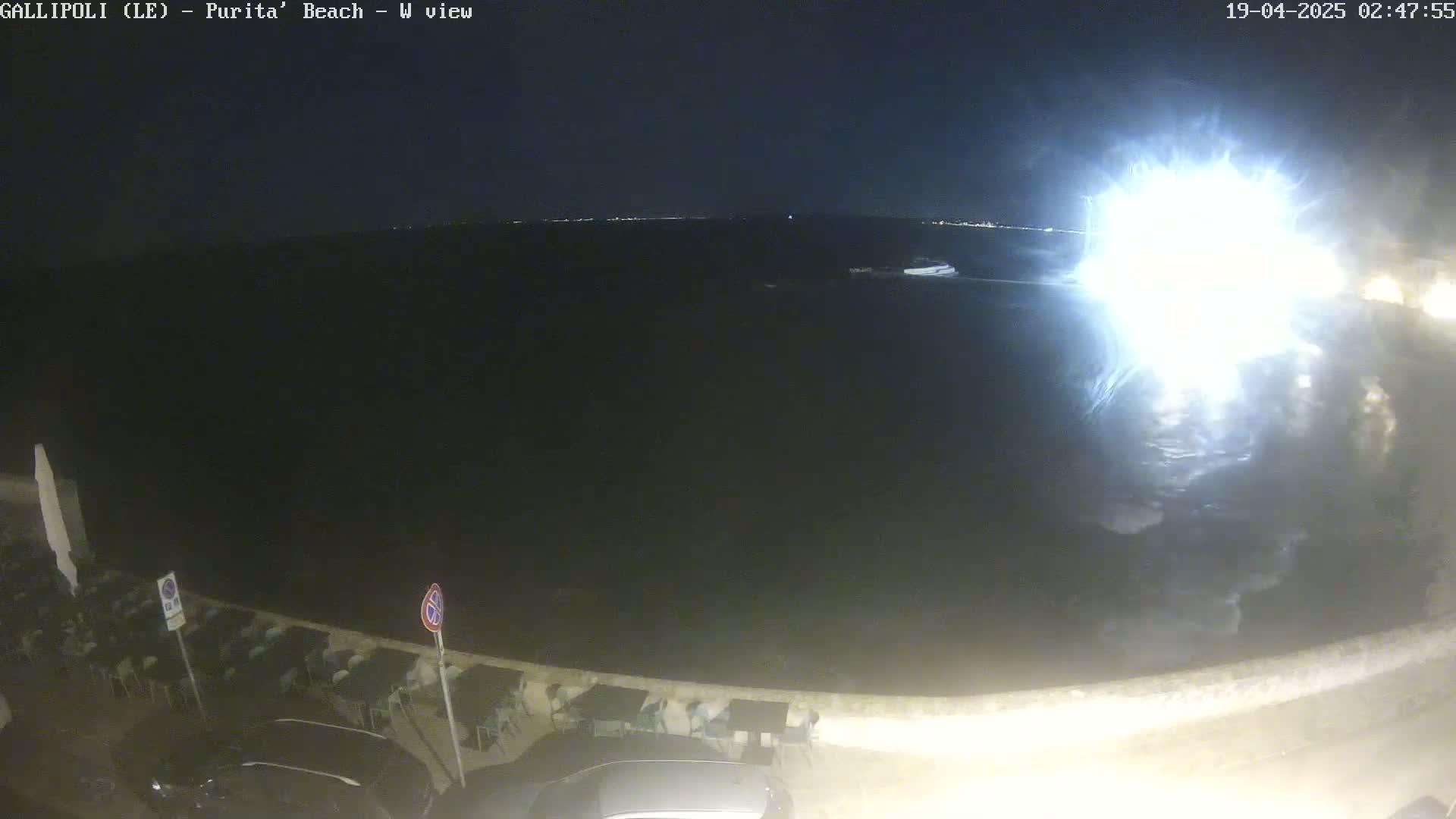 A nighttime view of a dark body of water with a bright light source in the distance, along a shoreline with parked cars and outdoor seating.