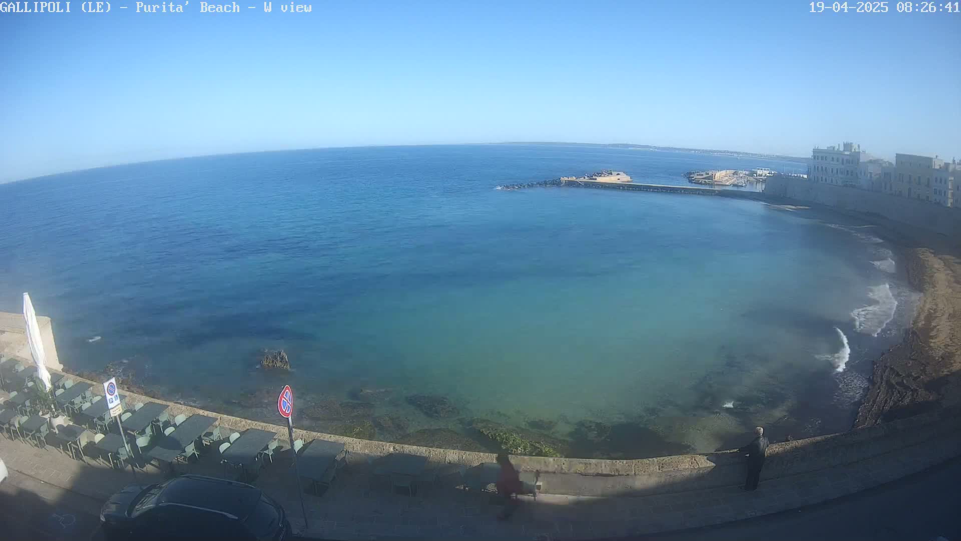 A clear, sunny day shows a calm, teal-colored ocean lapping a rocky shore next to a stone building and outdoor seating area with a few people present.