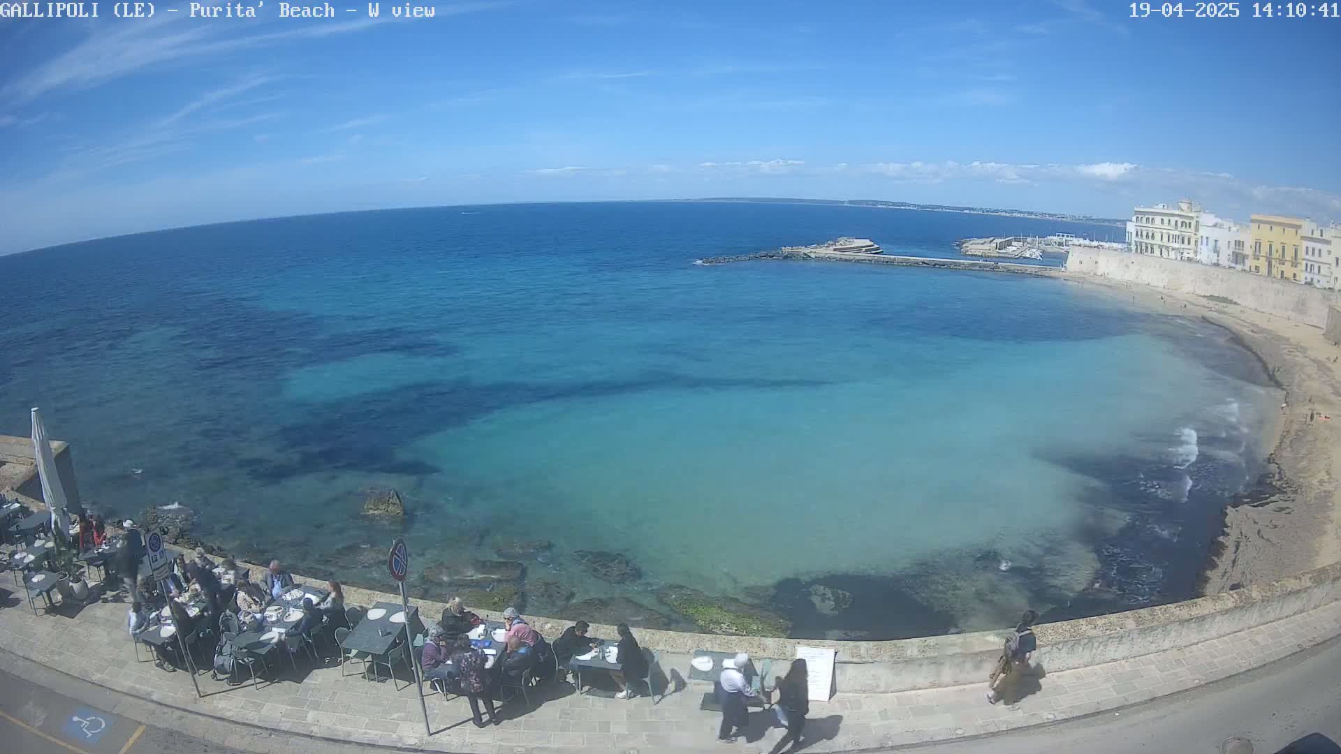 A seaside restaurant with patrons seated at tables overlooks a calm, clear turquoise bay on a sunny day.