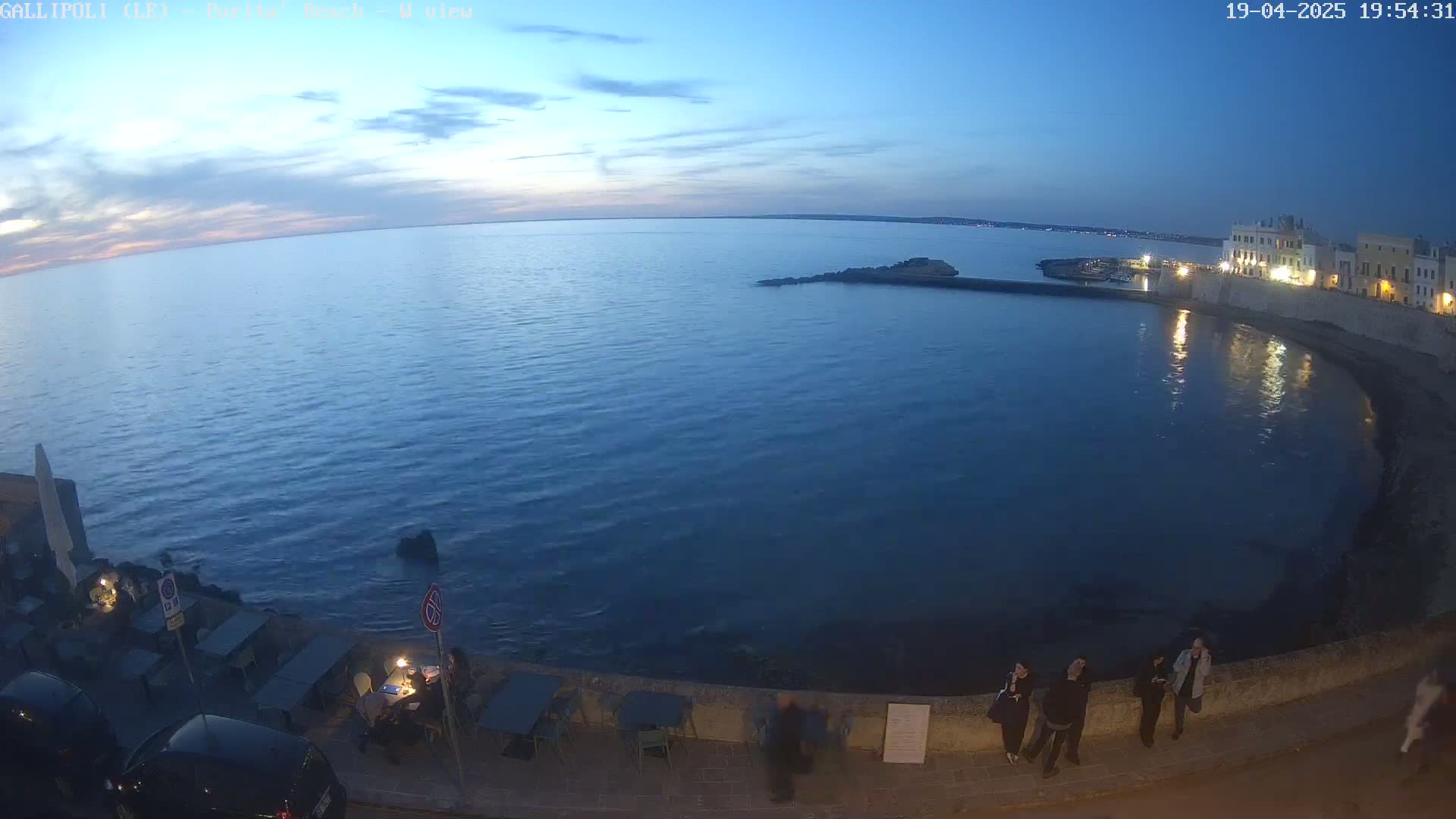 A dark-blue sea under a twilight sky curves around a waterfront area with people standing along a wall next to outdoor seating.