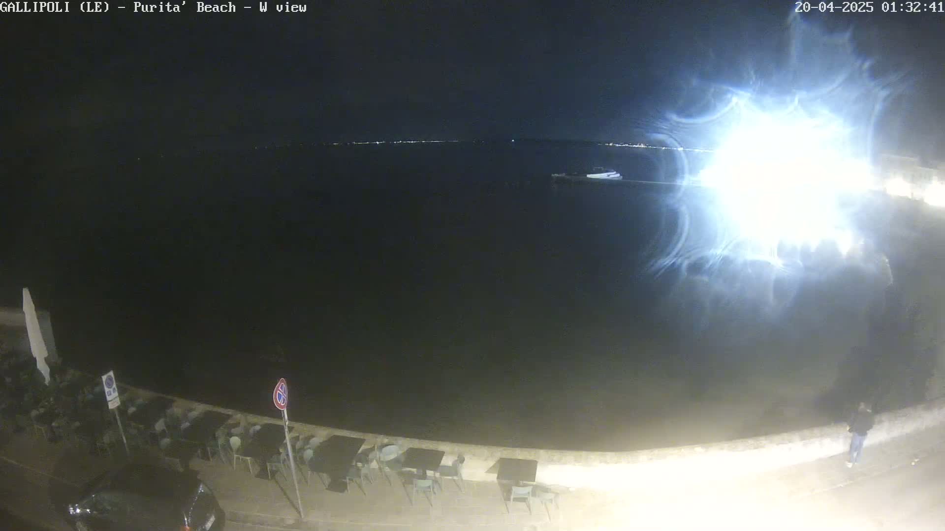A nighttime view of a dark body of water with a bright light reflecting on the surface,  a shoreline with outdoor seating, and a person standing near the water's edge.