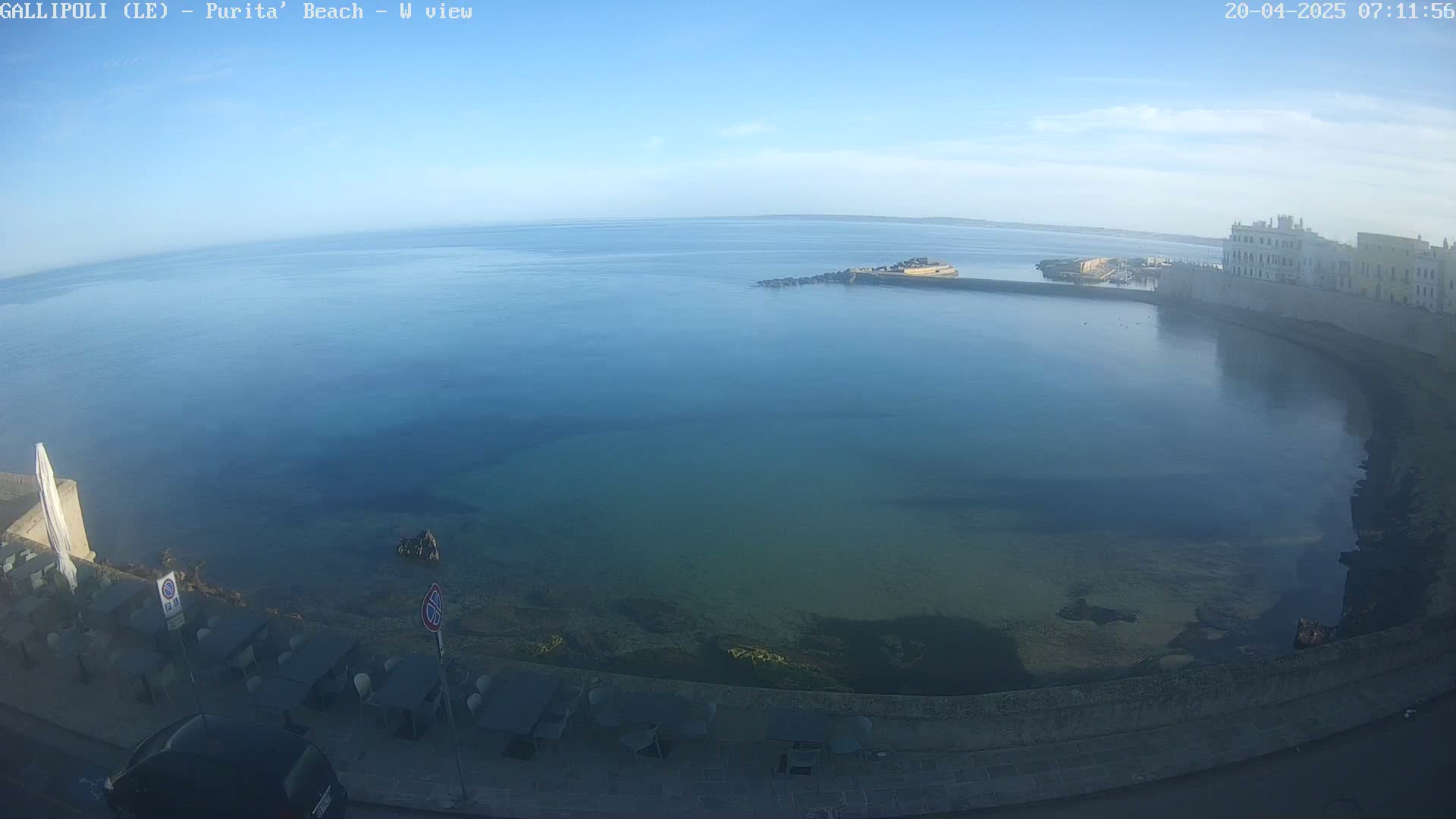 A clear, calm sea laps a stone shoreline with buildings and outdoor seating visible under a mostly clear sky.