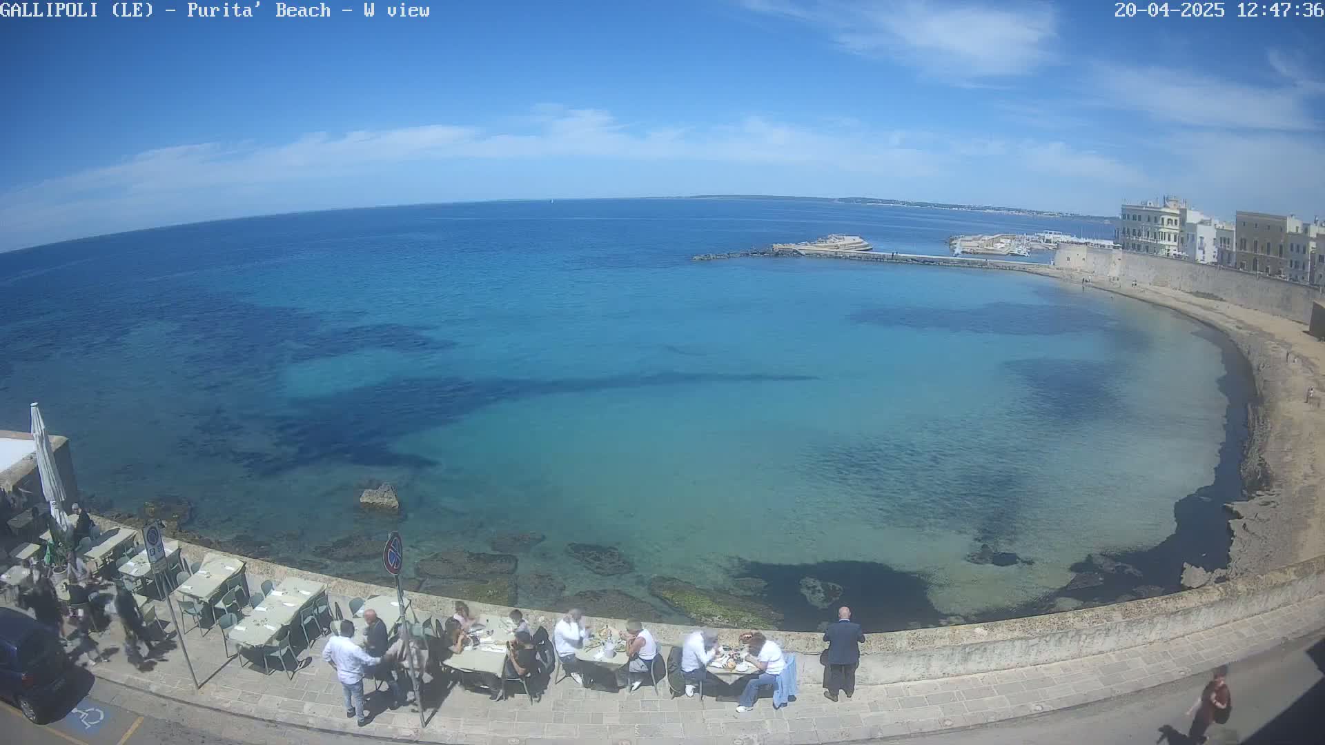 A sunny day shows a clear, turquoise-blue bay with people dining outdoors at a seaside restaurant next to a stone wall.