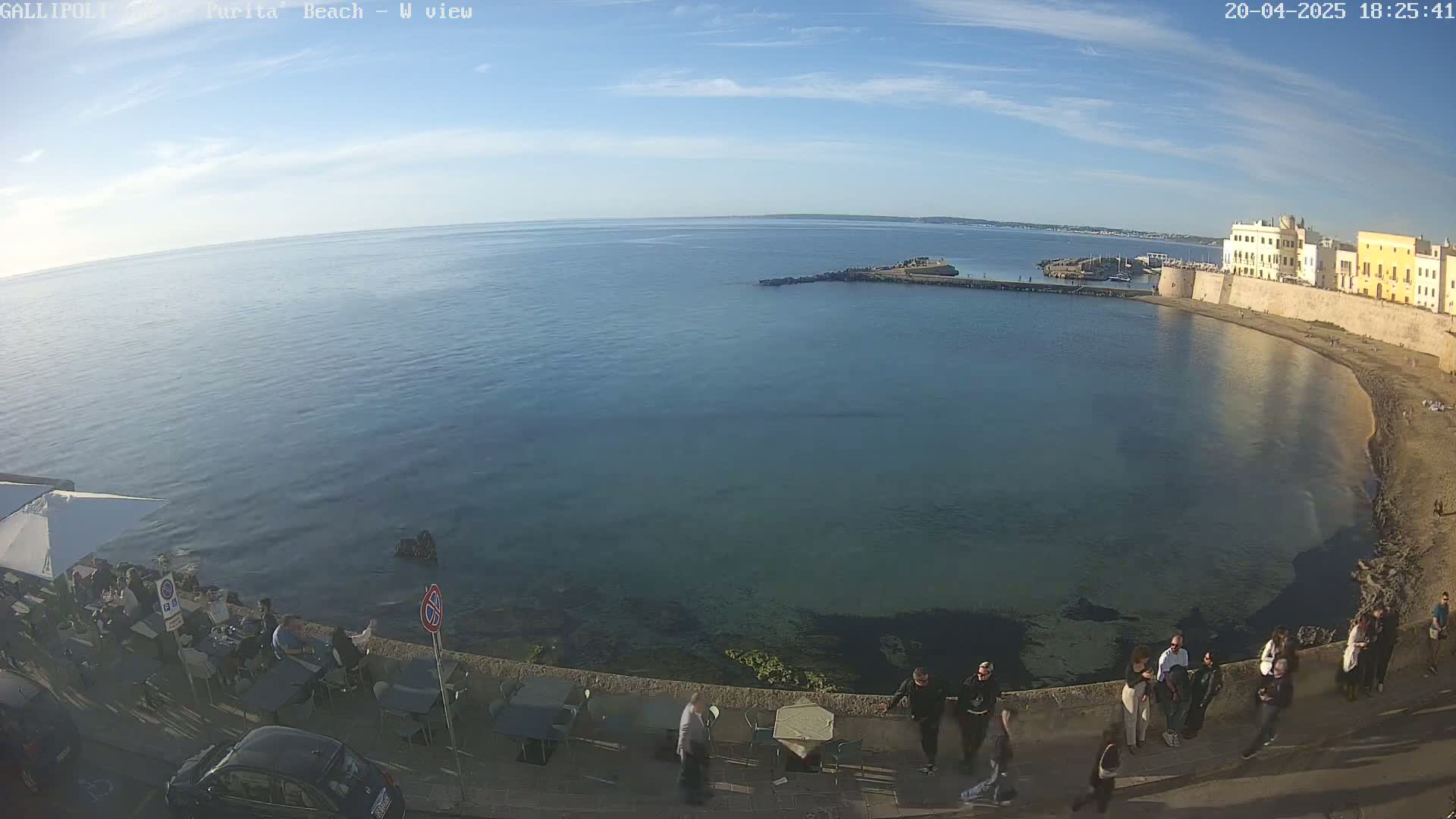 A mostly sunny day overlooks a calm, blue-green sea, a sandy beach, and a stone waterfront lined with buildings and people enjoying an outdoor setting.