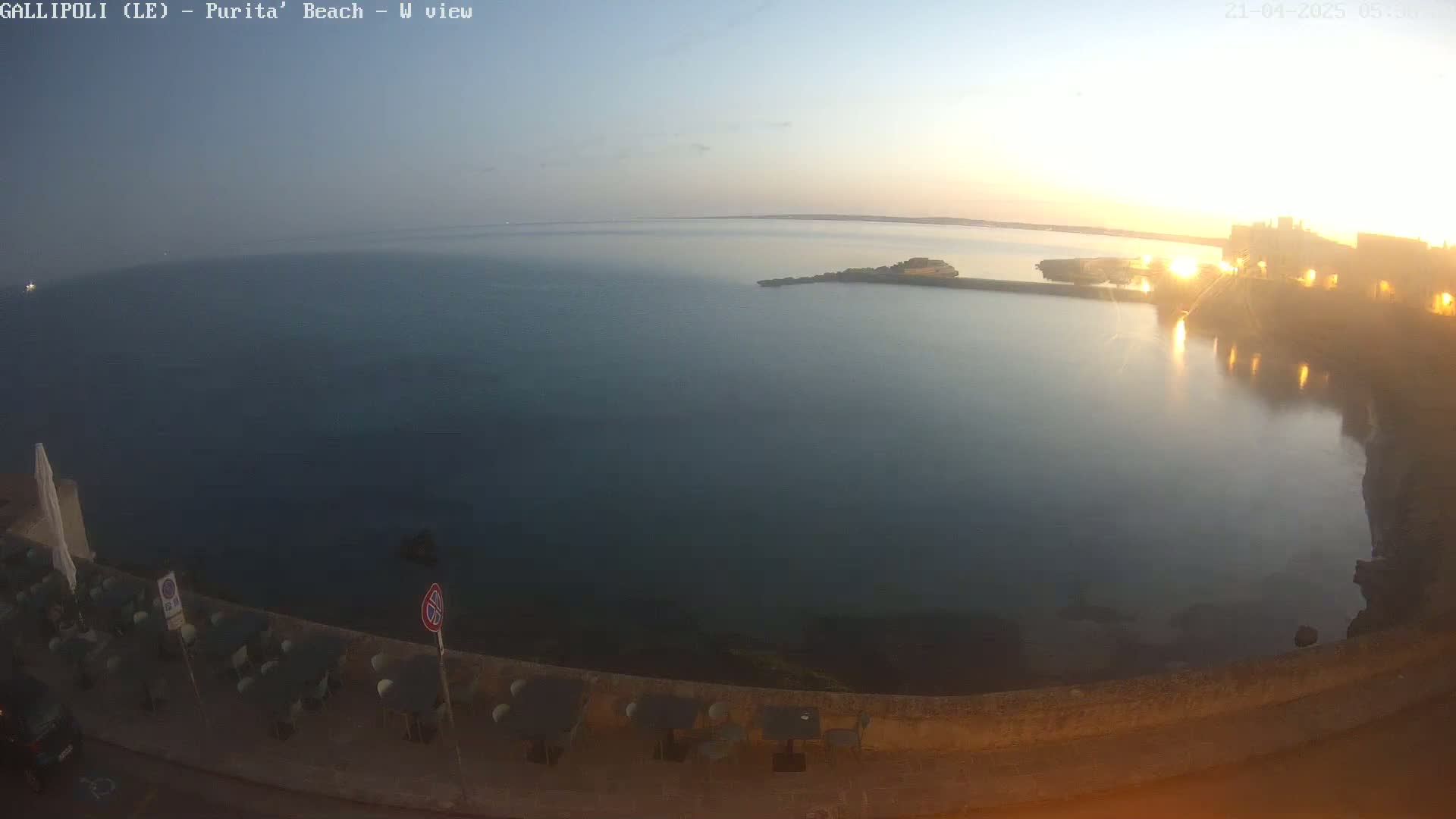 A calm, dark-blue sea at sunrise is seen from an elevated viewpoint overlooking a stone wall with outdoor seating and a partially lit town in the distance.
