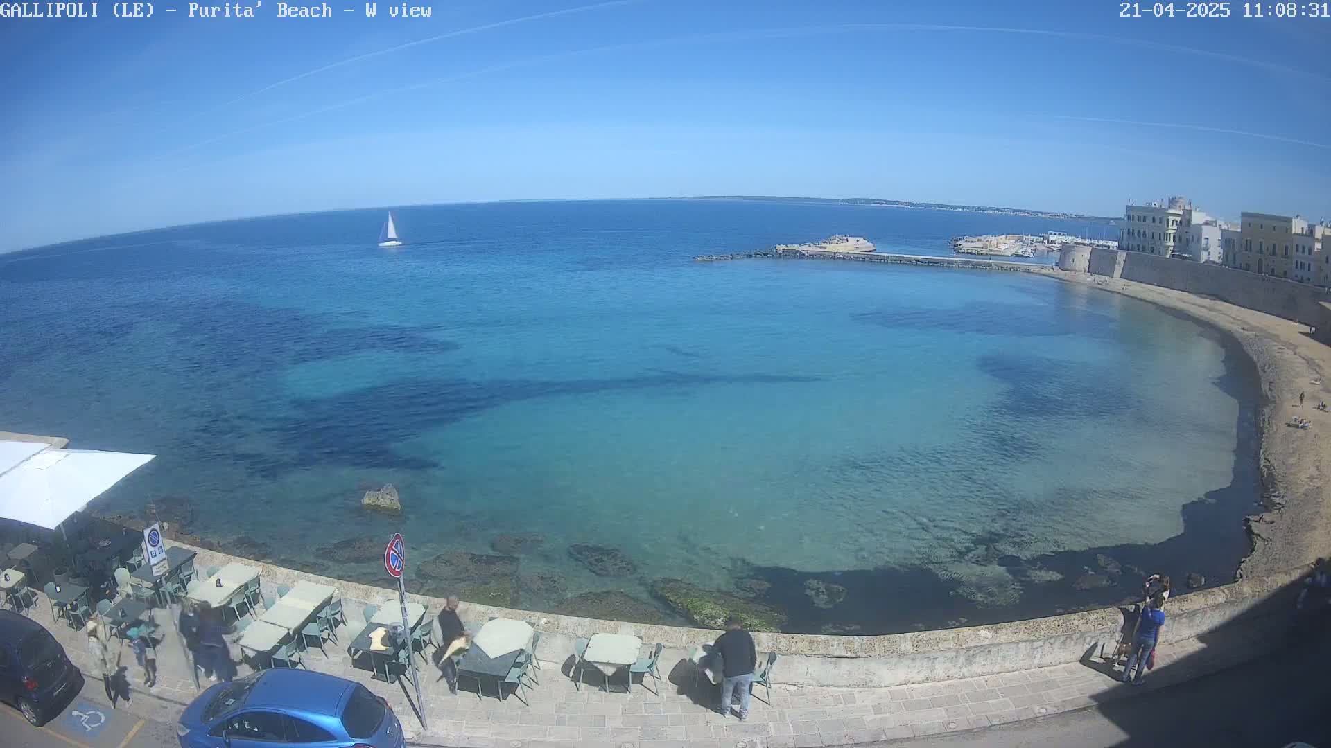 A clear, sunny day reveals a calm, turquoise bay with a small sailboat in the distance, a seaside restaurant with outdoor seating, and a stone wall bordering a sandy beach.