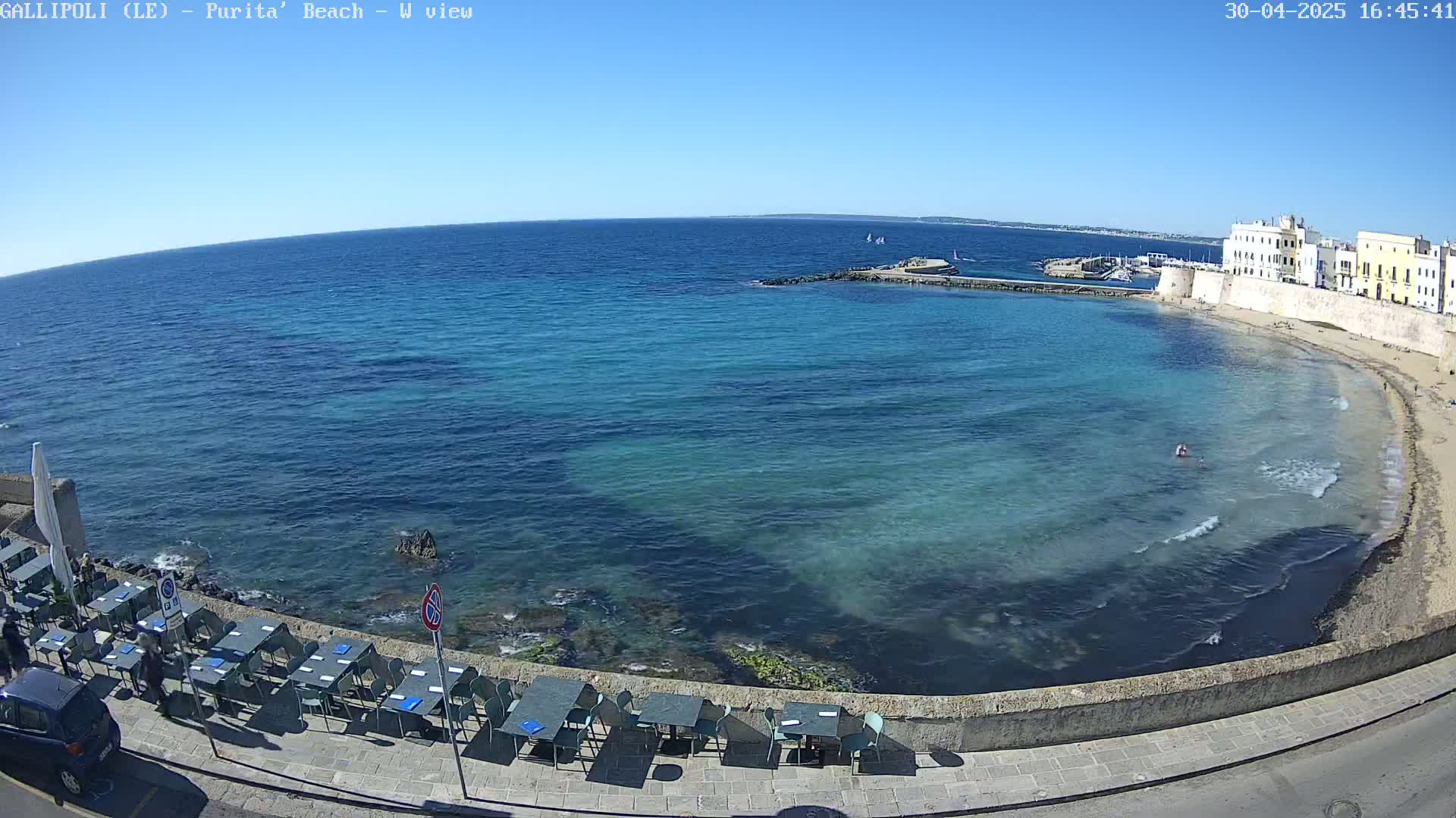 A clear, sunny day overlooks a calm, turquoise ocean lapping a sandy beach next to a stone wall and seaside restaurant with outdoor seating.