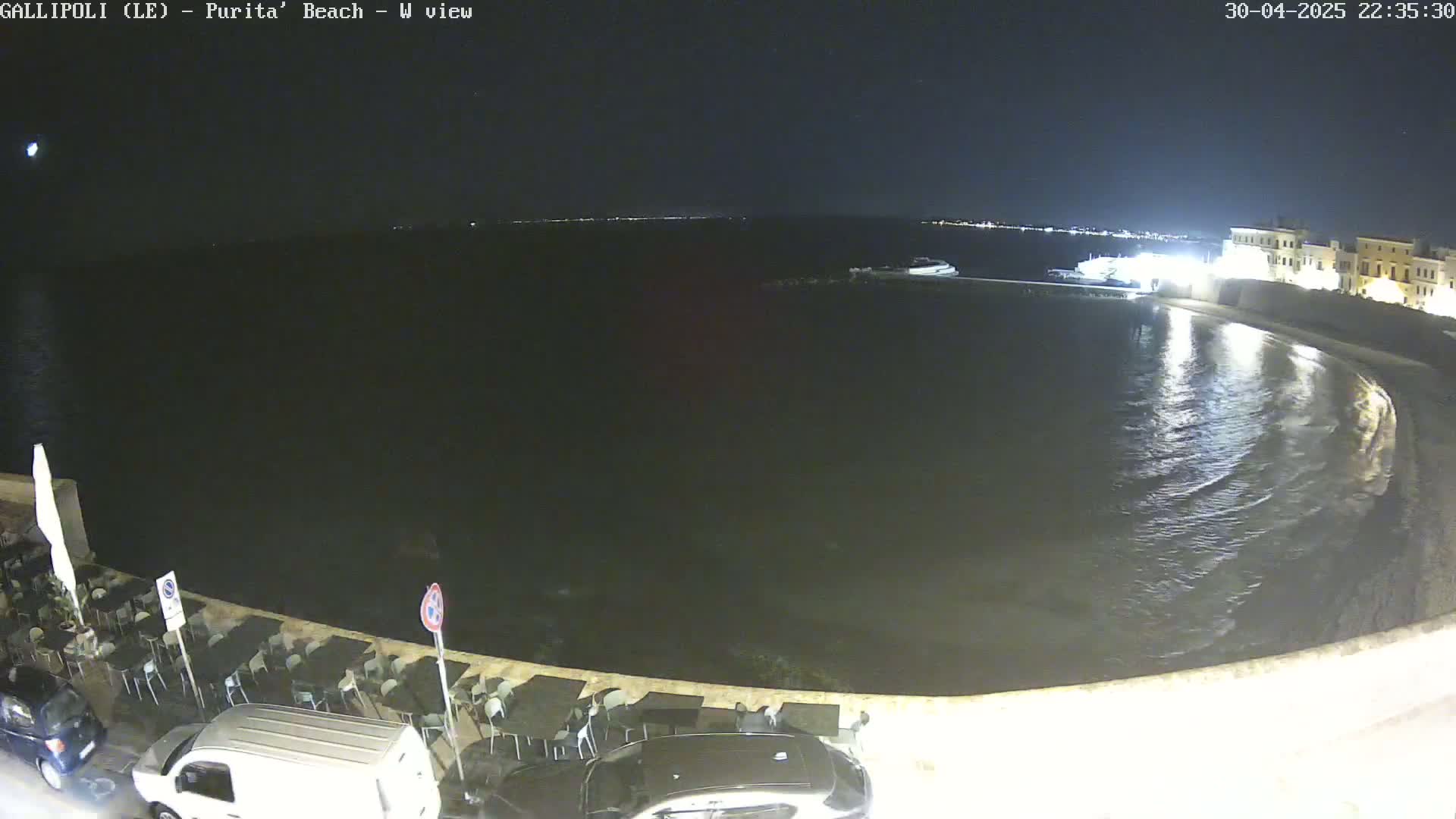 A nighttime view of a calm, dark body of water next to a shoreline with parked cars and outdoor seating.