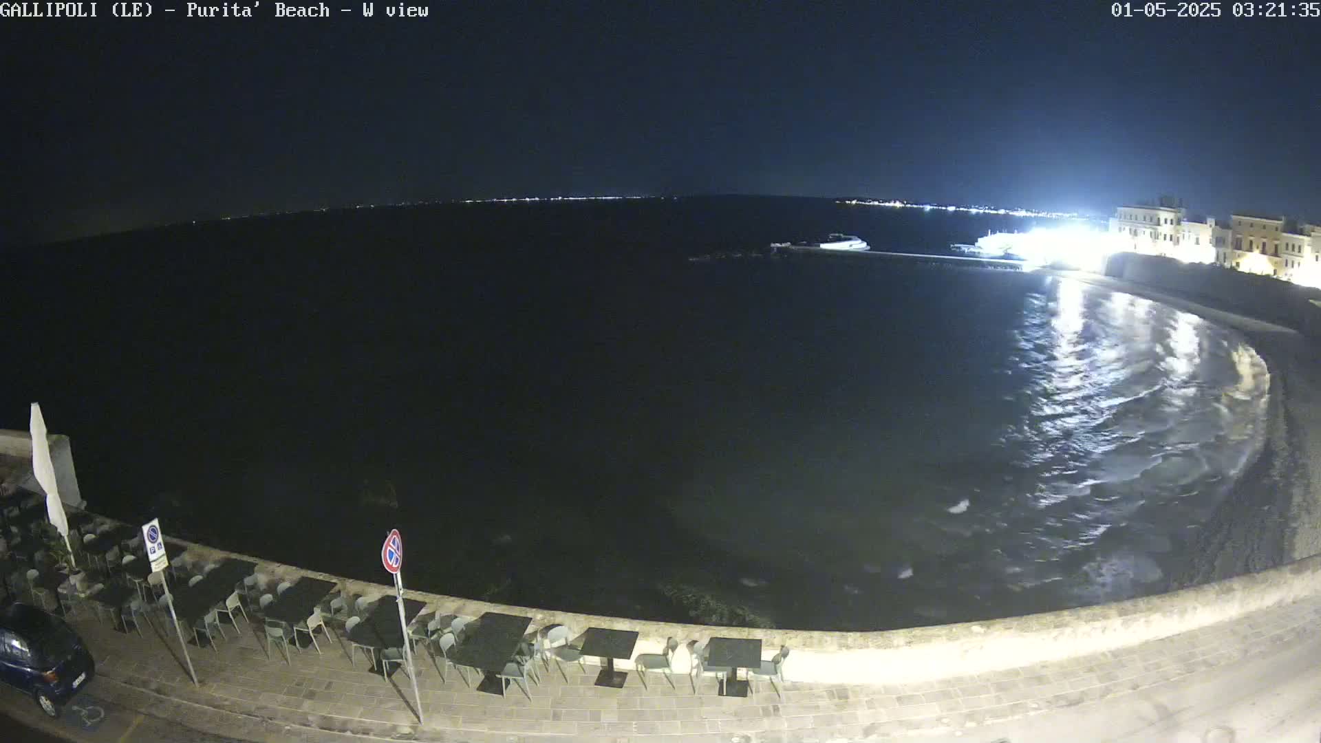 A nighttime view of a calm sea lapping at a stone shoreline, with empty outdoor seating visible in the foreground.