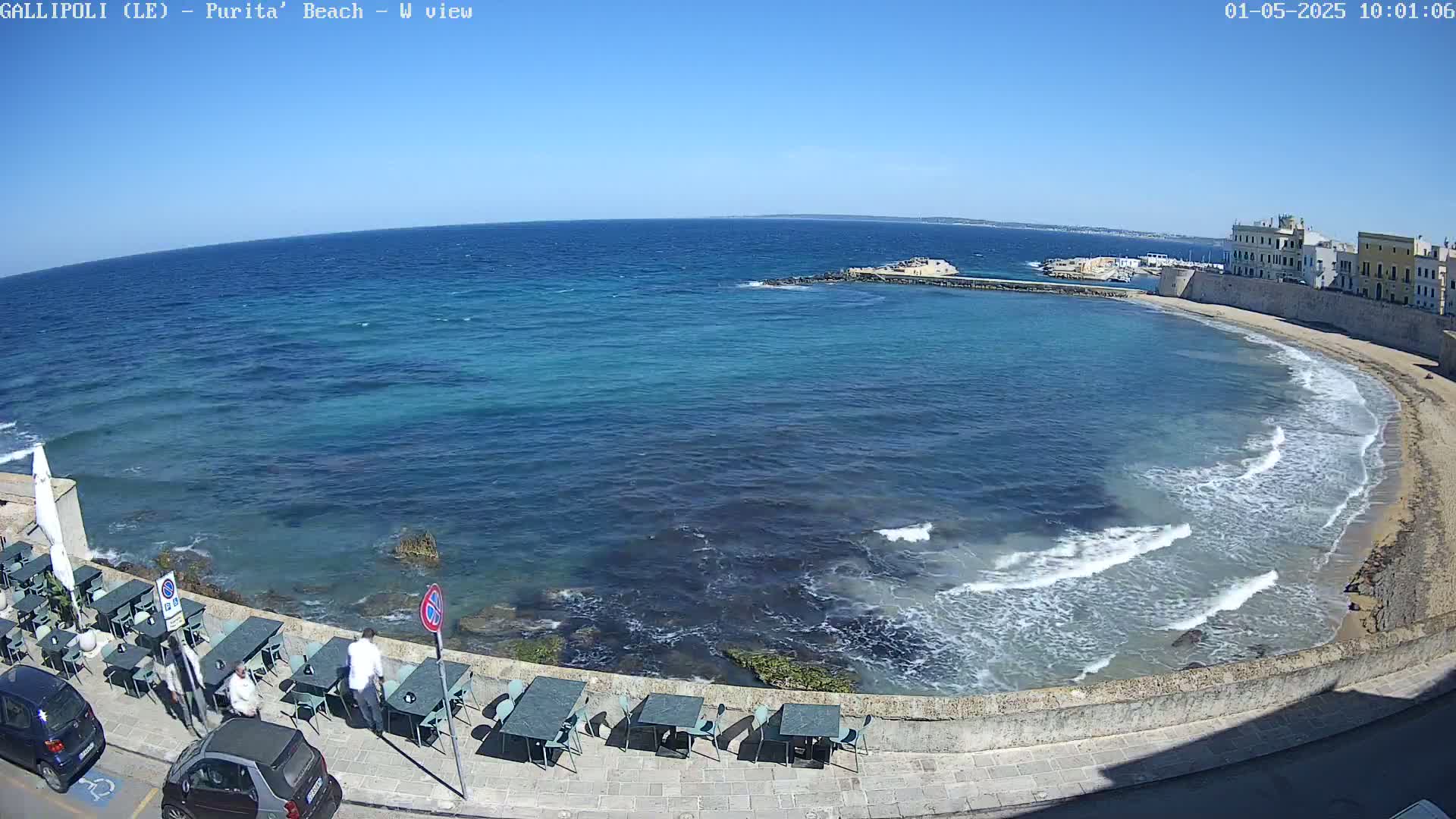A clear, sunny day reveals a calm, blue sea lapping a sandy beach, with a stone wall and outdoor seating lining the shore.