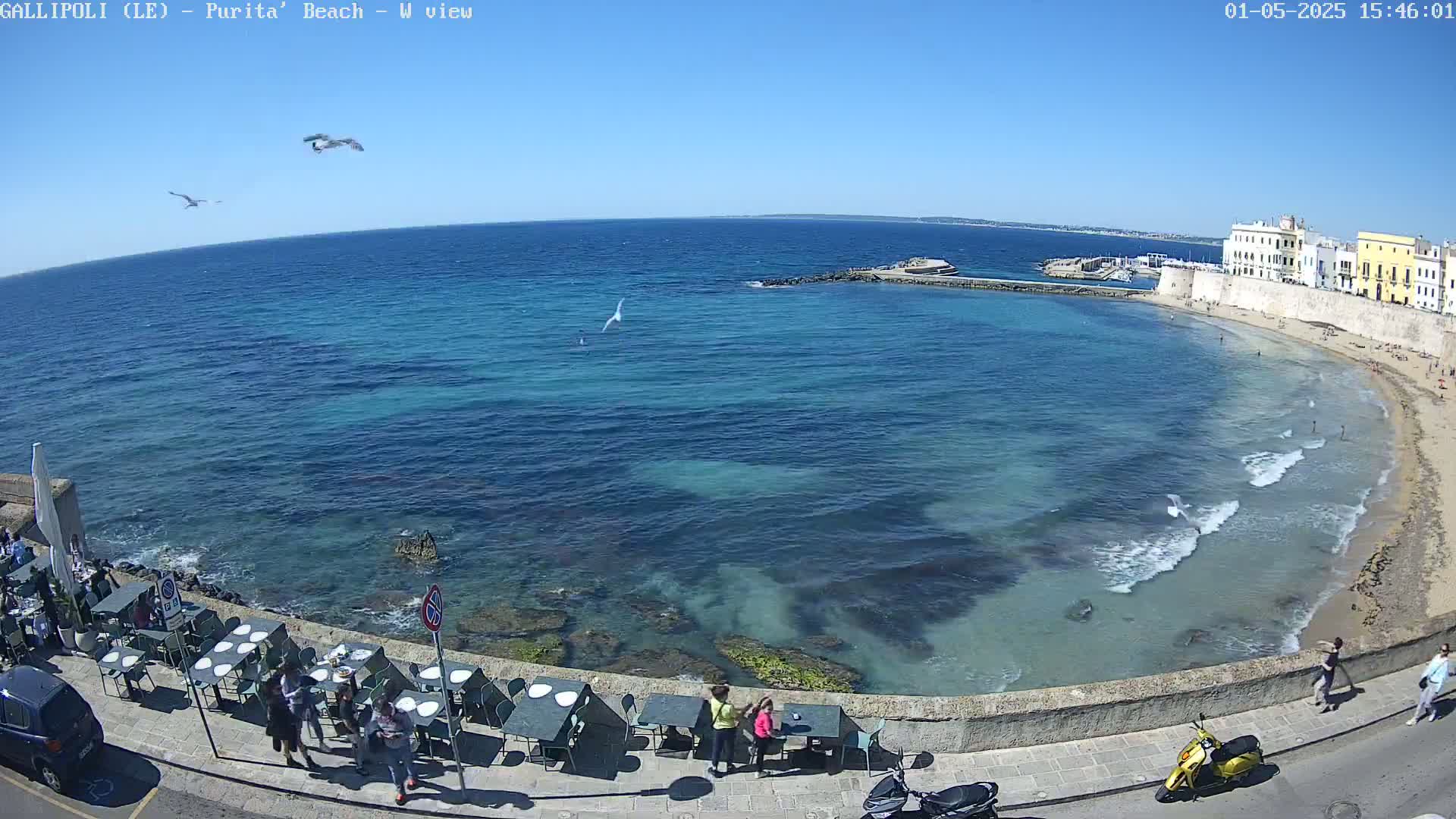A seaside town with a sandy beach and clear blue water under a bright sunny sky is seen from an elevated viewpoint, with several people strolling along the shore and seated at outdoor tables.