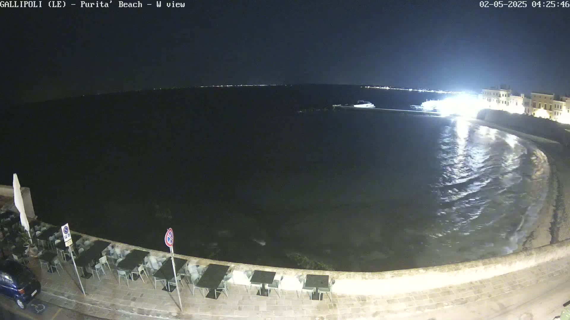 A nighttime seaside view shows gently lapping waves on a dark beach with outdoor tables and chairs at a restaurant next to a building, under a clear sky.
