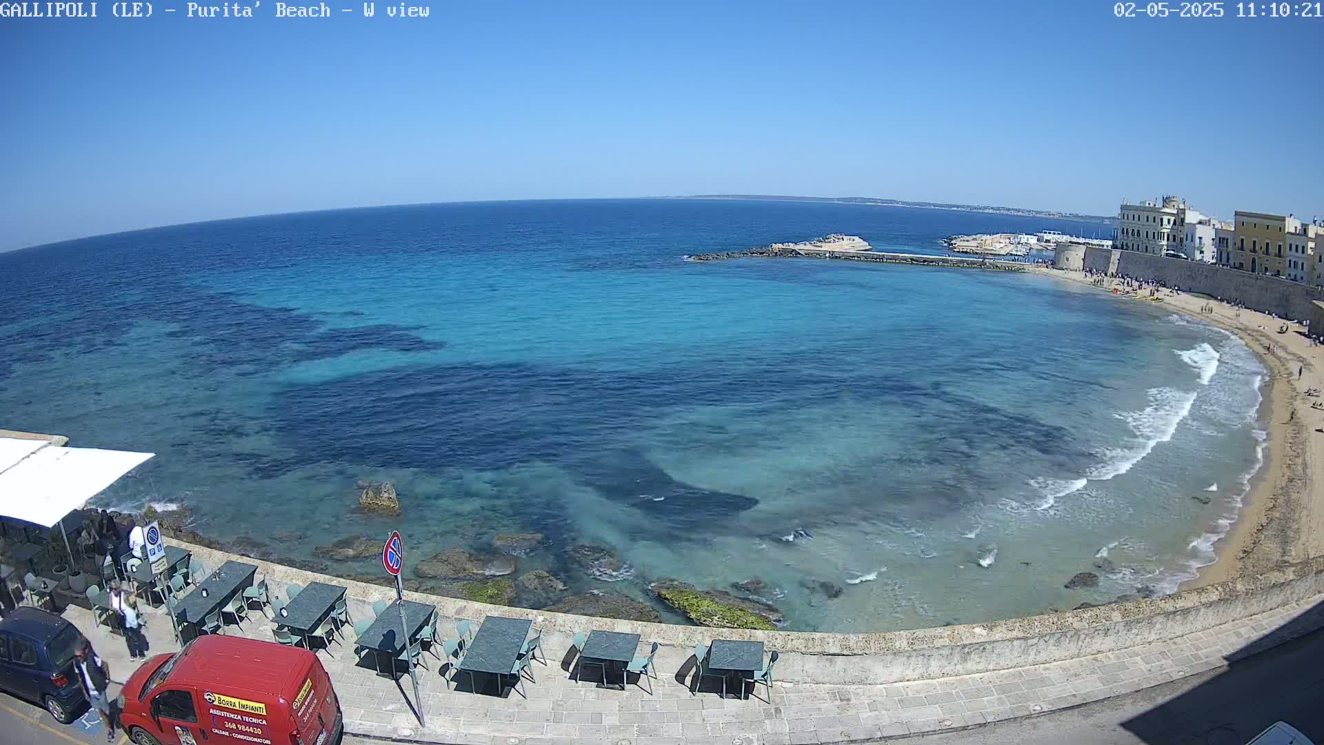 A clear, sunny day shows a beach with turquoise water lapping gently against the shore, featuring outdoor seating at a seaside restaurant and a small town visible in the distance.